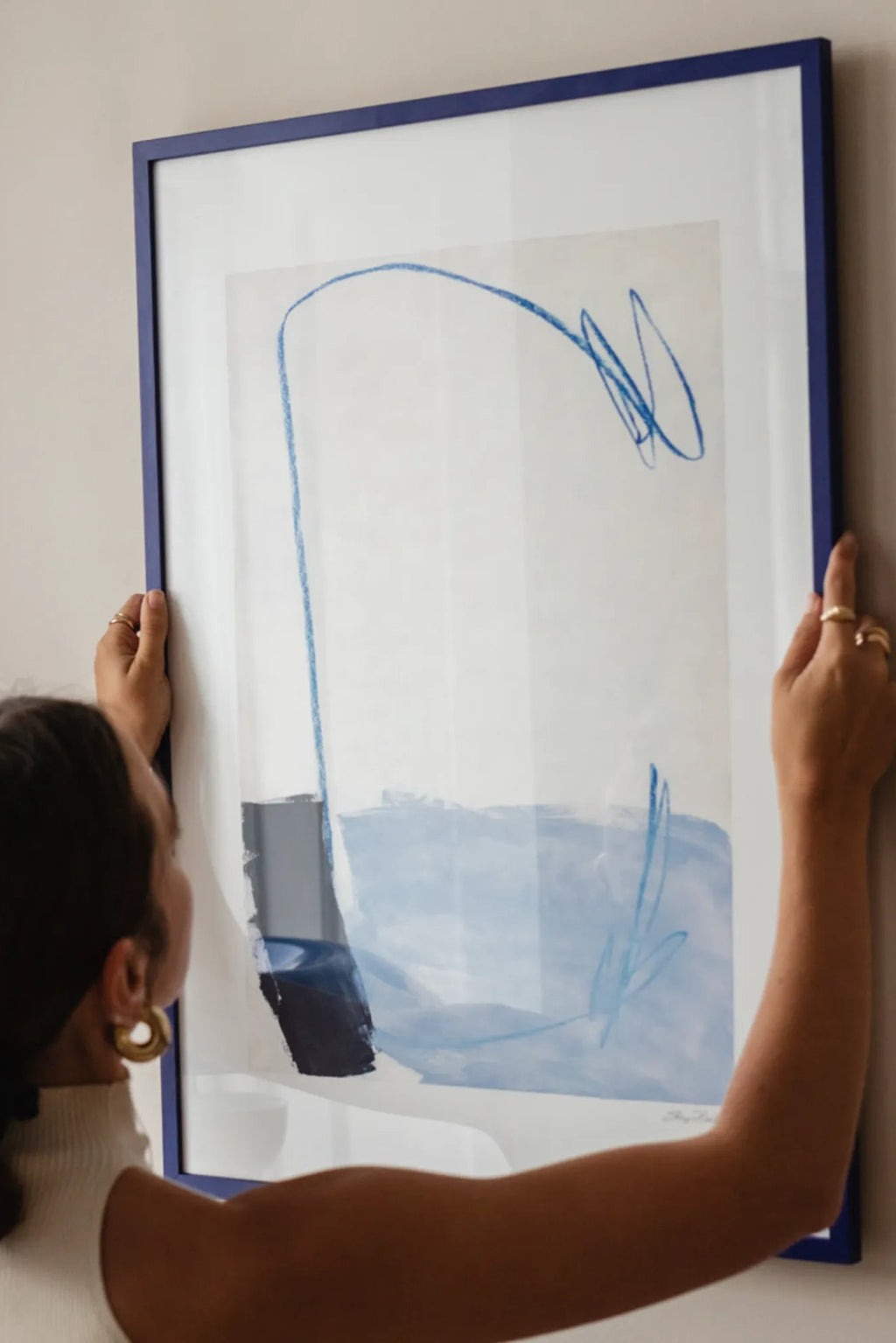 The Poster Club Kevät Saapuu, an abstract art print featuring energetic cobalt blue crayon-like lines and soft light blue watercolor washes against a crisp white background, accented with a dark navy block. This contemporary framed poster is elegantly presented with a wide white mat and a bold matte royal blue frame. Perfect as modern wall art for a minimalist living room or a vibrant addition to a gallery wall in Scandinavian or contemporary interiors.