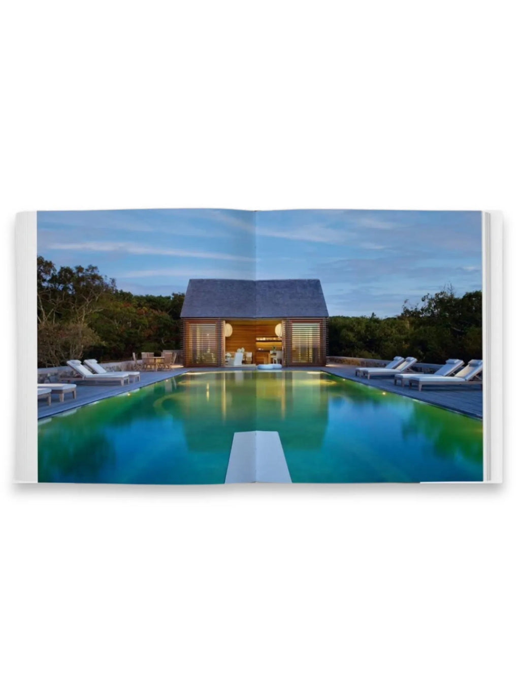 Book Shingle and Stone, an open coffee table book displaying a stunning architectural photograph of a modern, shingled pool house with wooden slats, illuminated interior, and a vibrant turquoise swimming pool surrounded by lounge chairs and lush greenery under a dusky sky. This premium design book provides inspiration for architects, interior designers, and homeowners interested in contemporary coastal living.