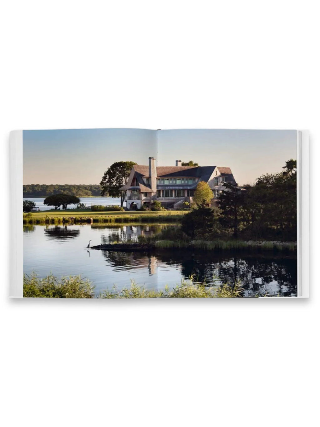 Book Shingle and Stone, an open book displaying a stunning full-bleed photograph of a luxurious shingle-style home nestled beside a tranquil lake with calm water reflecting the surrounding trees and clear sky. This architecture and design book is perfect for interior design enthusiasts and homeowners seeking inspiration for elegant, waterfront properties.