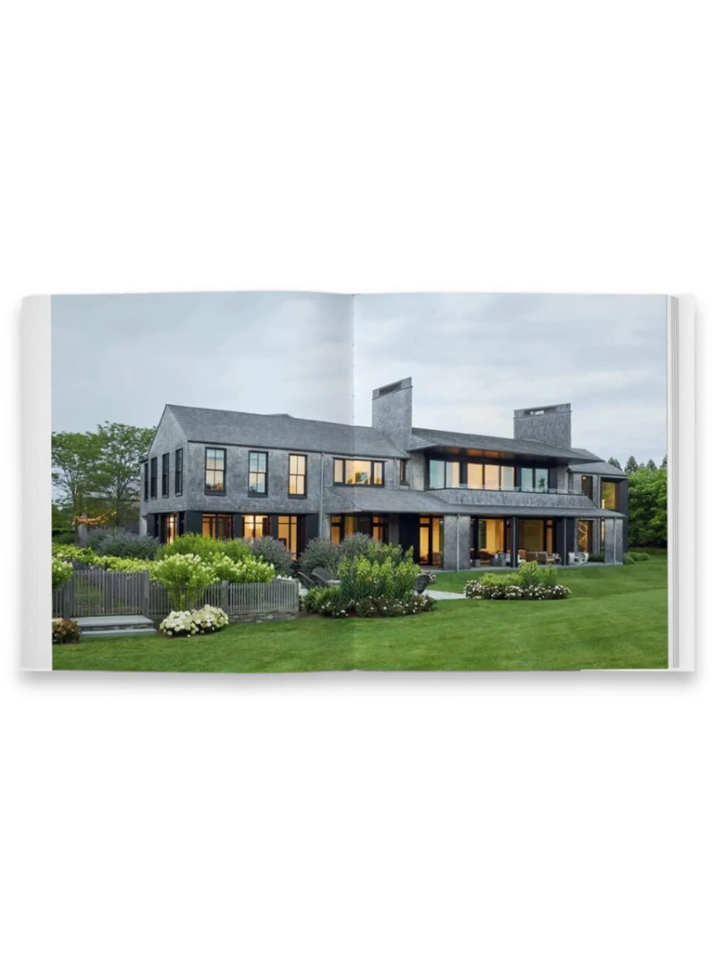 Book Shingle and Stone, an open coffee table book featuring a large, grey shingle-style modern luxury home with black window frames, expansive green lawns, and lush landscaping, under an overcast sky. This architectural design book is perfect for interior designers, architects, and homeowners seeking inspiration for elegant, contemporary residences.