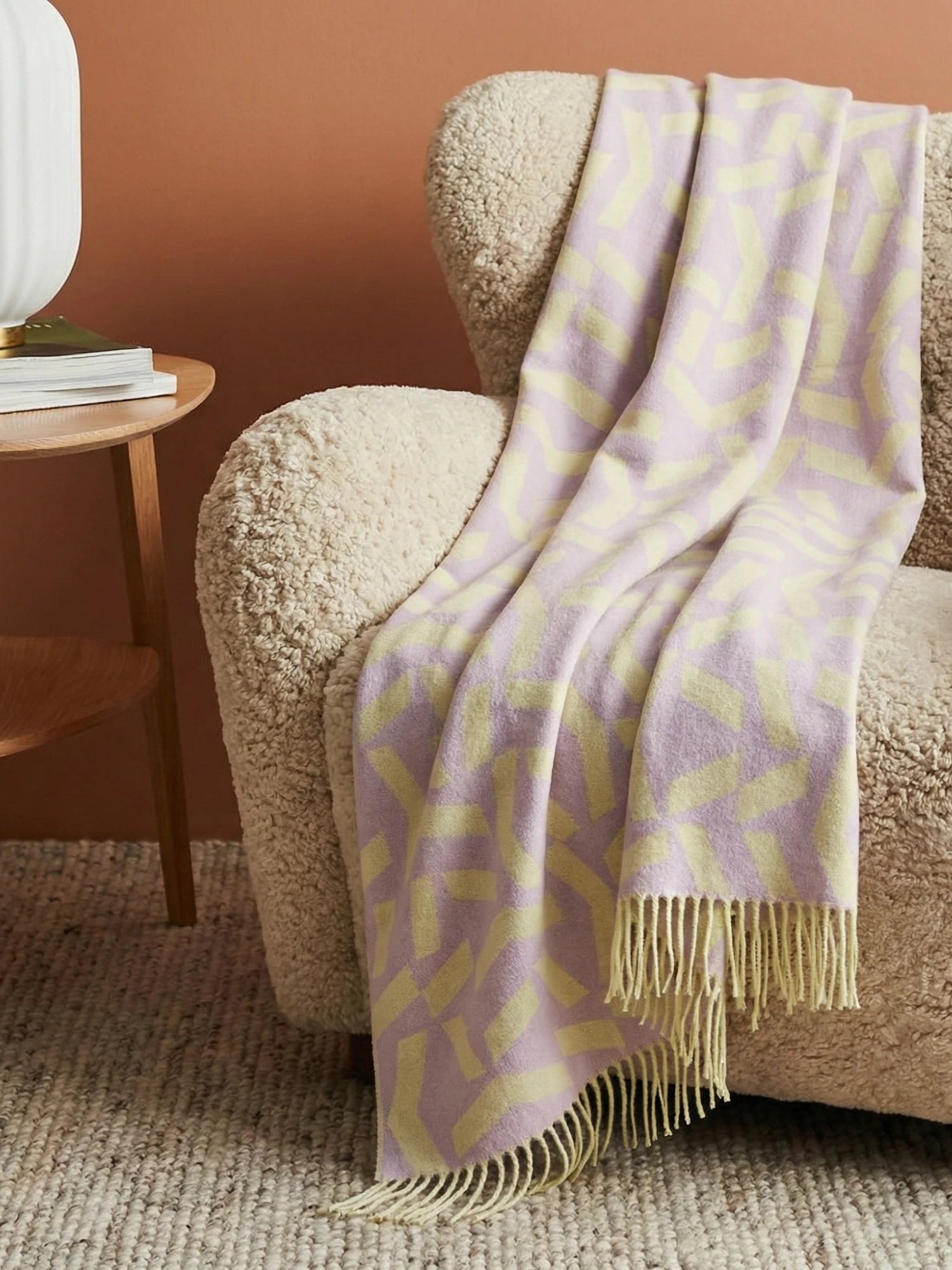 Lavender and yellow patterned throw blanket draped over a textured beige lounge chair against a brown wall.