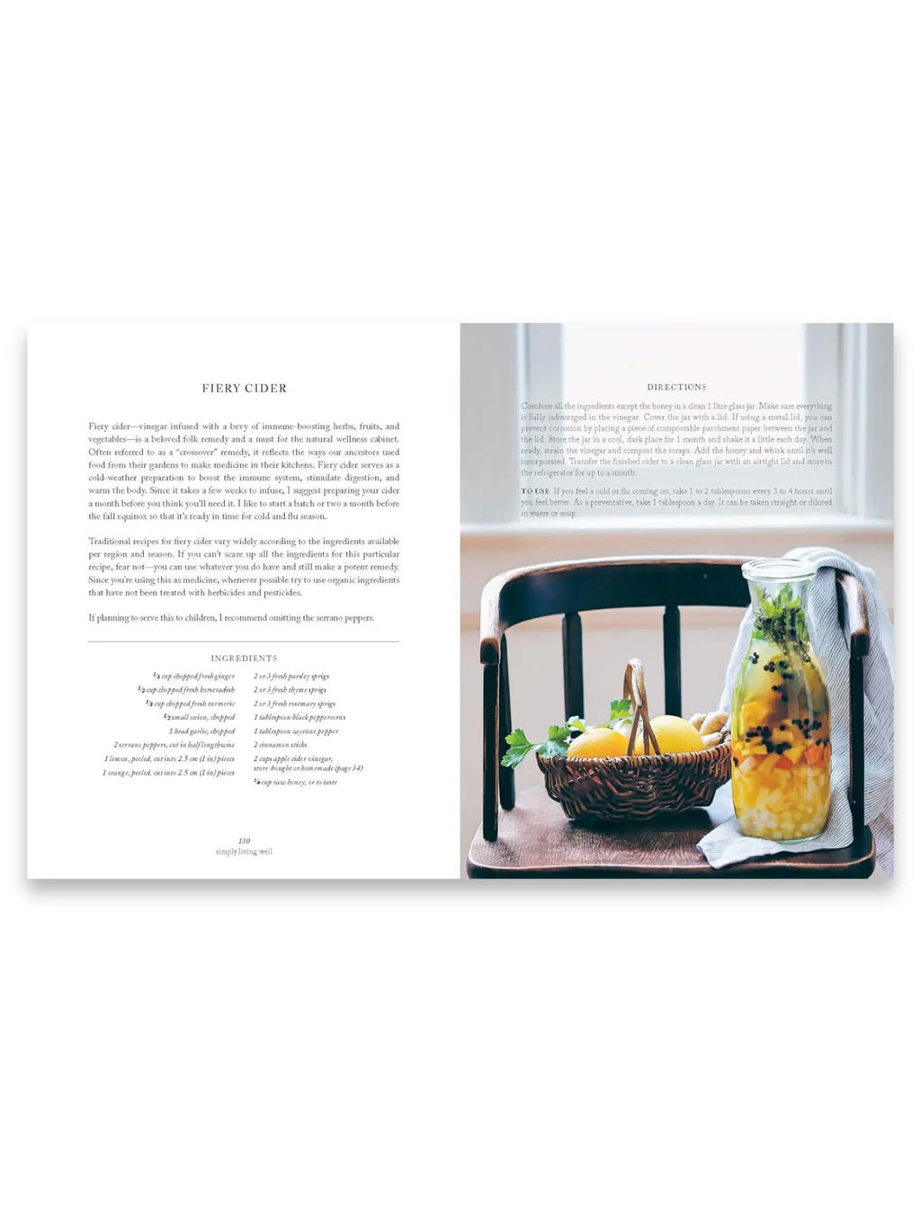 Book Simply Living Well, an open cookbook page displaying a "Fiery Cider" recipe with vibrant ingredients like lemons, ginger, and herbs in a clear glass jar on a rustic wooden chair, alongside a woven basket of fresh produce. This comprehensive natural wellness guide is ideal for home cooks and health-conscious individuals seeking wholesome remedies and sustainable living practices.