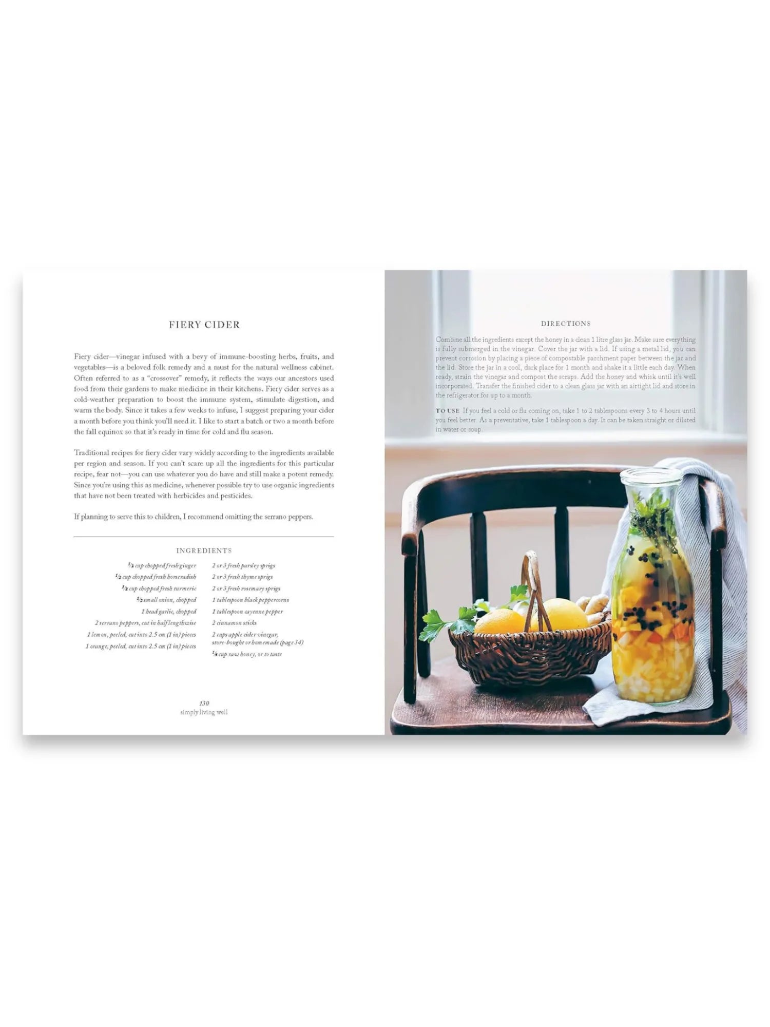 Book Simply Living Well, an open cookbook page displaying a "Fiery Cider" recipe with vibrant ingredients like lemons, ginger, and herbs in a clear glass jar on a rustic wooden chair, alongside a woven basket of fresh produce. This comprehensive natural wellness guide is ideal for home cooks and health-conscious individuals seeking wholesome remedies and sustainable living practices.