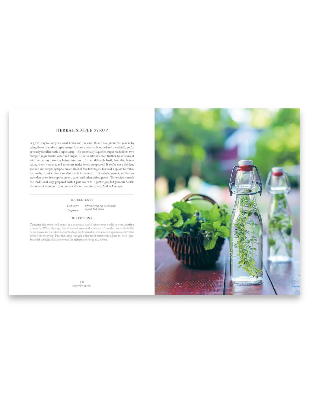 Book Simply Living Well, an open cookbook page displaying a "Herbal Simple Syrup" recipe with text on the left and a vibrant photo on the right. The image showcases two clear glass swing-top bottles filled with herbal simple syrup, featuring fresh green herbs submerged in liquid, alongside a rustic woven basket overflowing with fresh mint on a warm wooden table. This essential book offers practical, natural recipes for home chefs and wellness enthusiasts.
