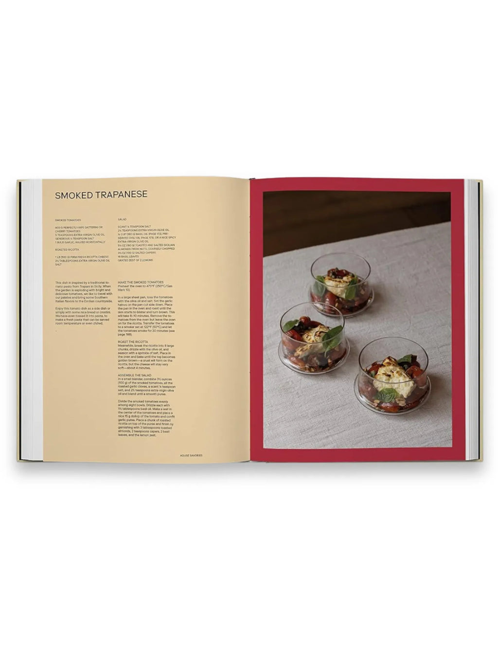 Book Slow Food, Fast Cars, an open cookbook displaying a "Smoked Trapanese" recipe with detailed text on the left page and a vibrant food photograph on the right. The image features three elegant glass bowls filled with cherry tomatoes, basil leaves, and creamy ricotta cheese, artfully arranged on a light gray linen tablecloth within a deep red border. This premium cookbook is perfect for home chefs and culinary enthusiasts seeking stylish, Italian-inspired recipes.