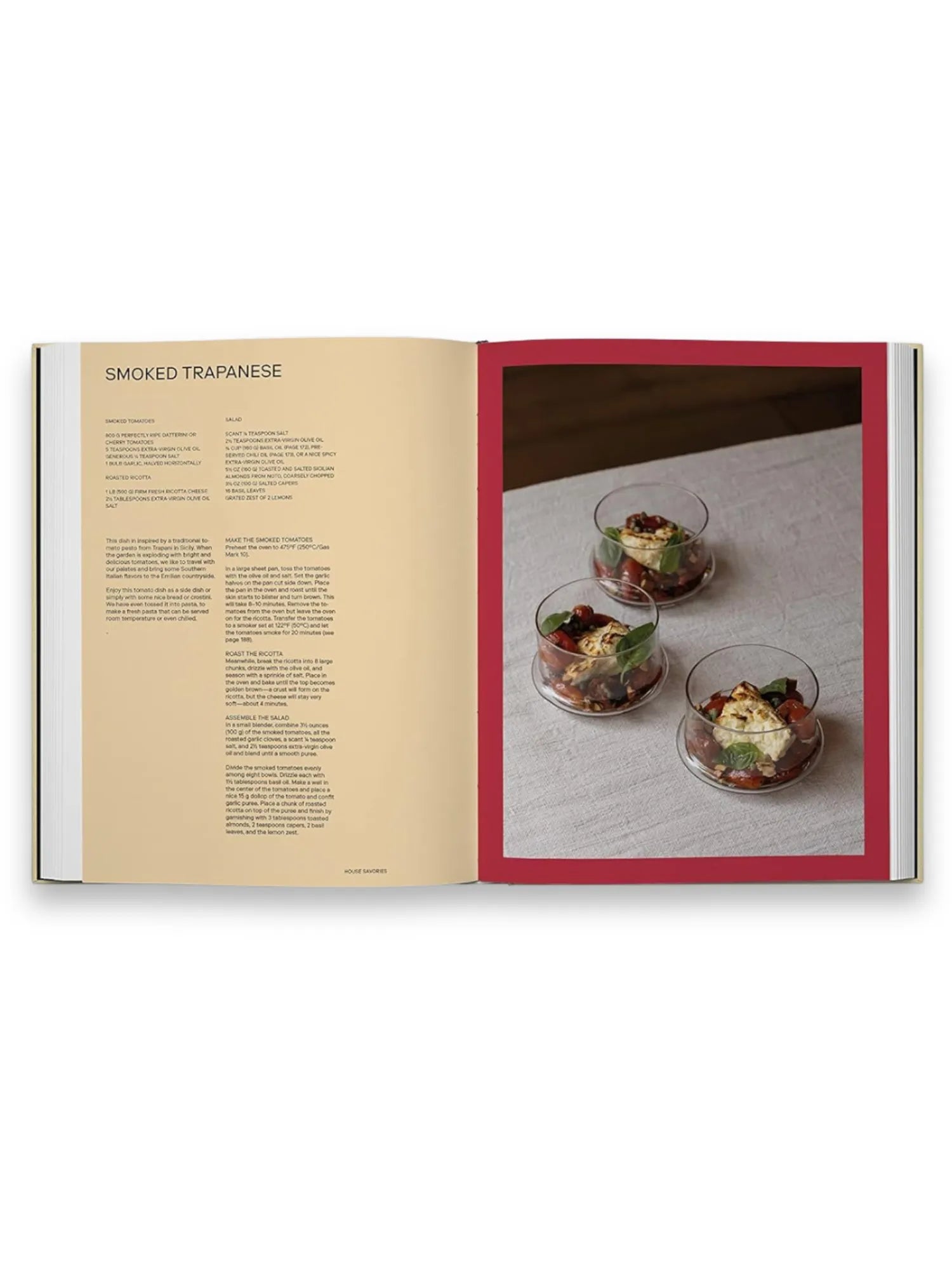 Book Slow Food, Fast Cars, an open cookbook displaying a "Smoked Trapanese" recipe with detailed text on the left page and a vibrant food photograph on the right. The image features three elegant glass bowls filled with cherry tomatoes, basil leaves, and creamy ricotta cheese, artfully arranged on a light gray linen tablecloth within a deep red border. This premium cookbook is perfect for home chefs and culinary enthusiasts seeking stylish, Italian-inspired recipes.