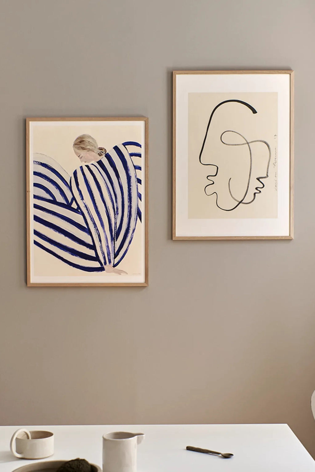 The Poster Club Blue Stripe At Concorde, an illustrative contemporary art print featuring a seated figure draped in bold, hand-painted deep blue and cream stripes, creating a dynamic visual flow. This minimalist paper art print is framed in a light natural oak frame, offering a clean, Scandinavian aesthetic. Perfect for modern gallery walls, adding a serene and stylish accent to contemporary living rooms or bedrooms.