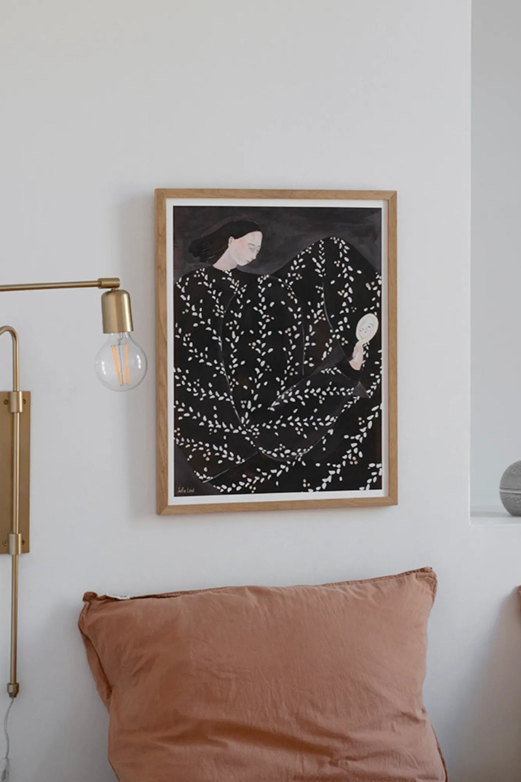 The Poster Club Mirror Mirror, a contemporary illustrative art print depicting a woman with dark hair in a black dress adorned with an organic white leaf pattern, holding a small white hand mirror. This framed art print is presented in a light natural oak frame with a crisp white mat. Ideal as minimalist wall art for a modern bedroom, living room, or gallery wall in Scandinavian or contemporary interiors.