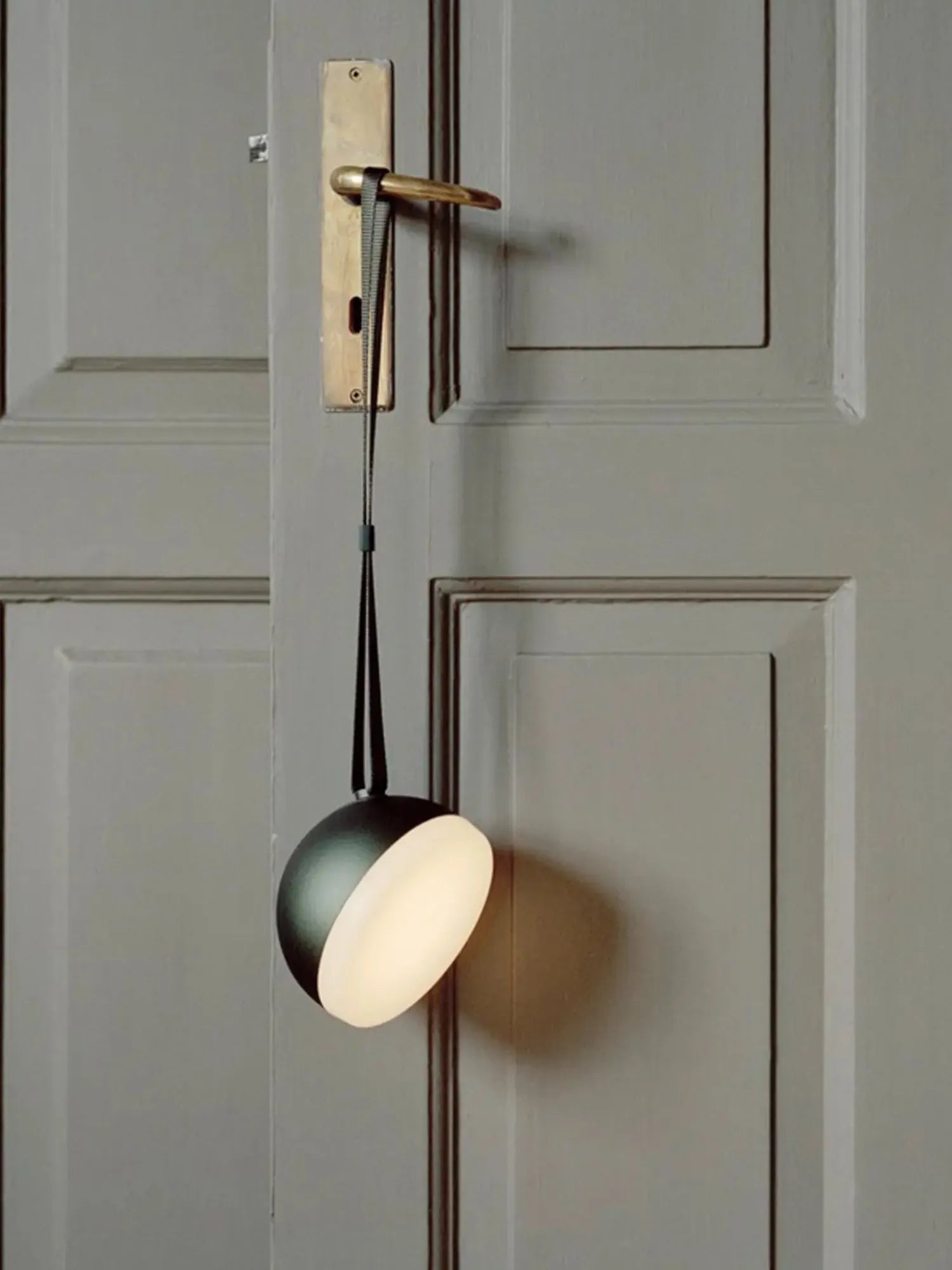 New Works Sphere Adventure Light, a black and white spherical portable lamp with a warm glow, hanging from a brass handle by a black strap. Ideal for chic portable lighting.