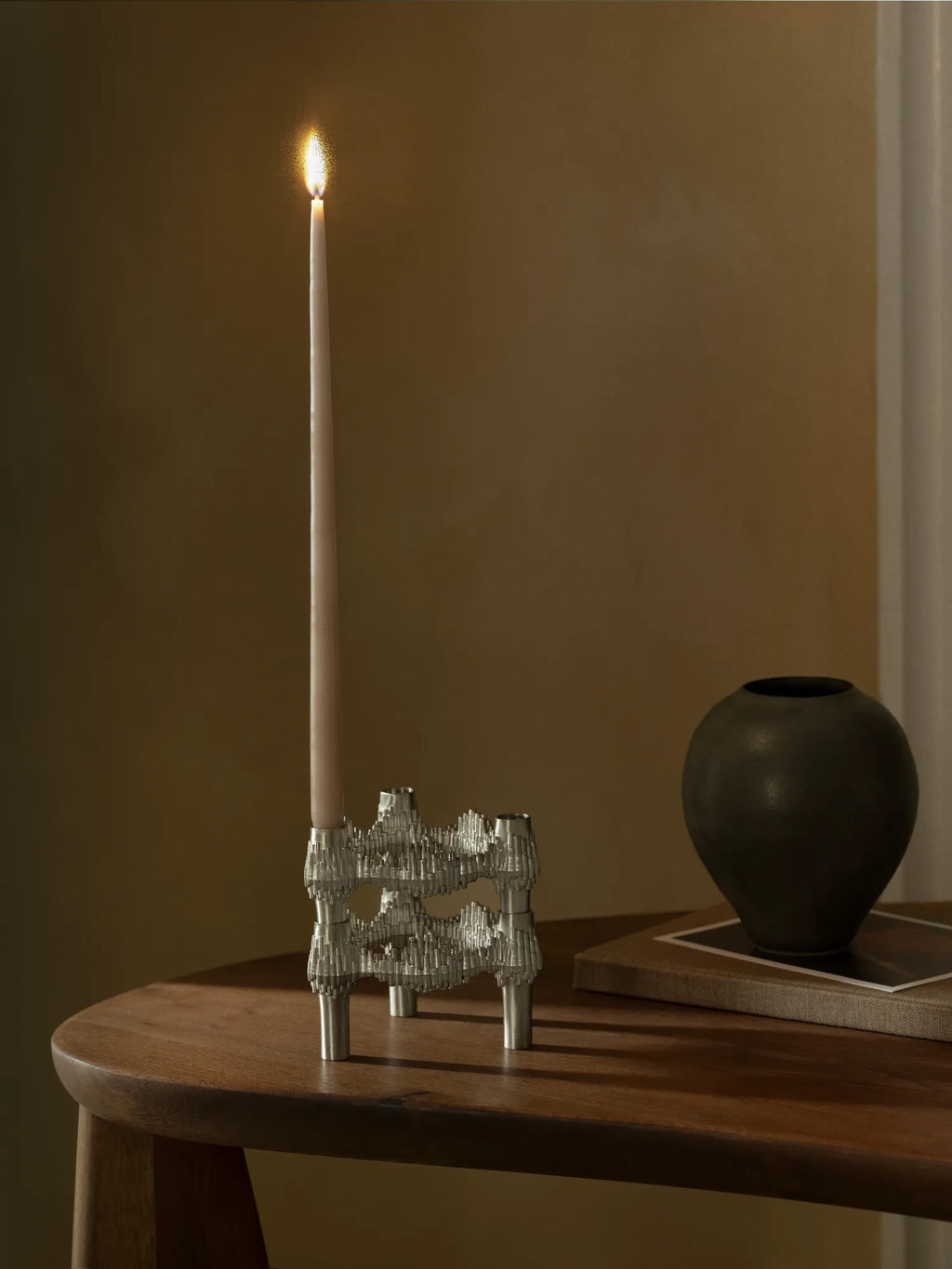 Stoff Nagel Quist Candle Holder, an iconic mid-century modern modular candle holder crafted from polished silver-toned metal with a distinctive brutalist, sculptural texture and interlocking design, currently holding a single cream taper candle. Perfect as a contemporary dining table centerpiece or a striking shelf styling accent for design-conscious interiors.