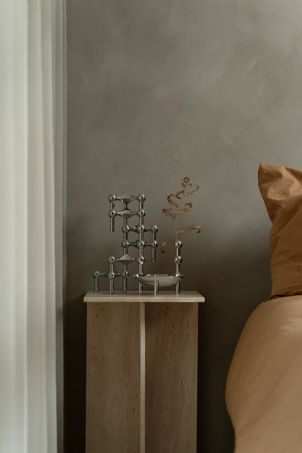 Stoff Nagel Candle Holder in a polished chrome finish, assembled into a compact, multi-level design, holding a dried botanical accent, resting on a sleek marble-topped side table beside a cozy bed.