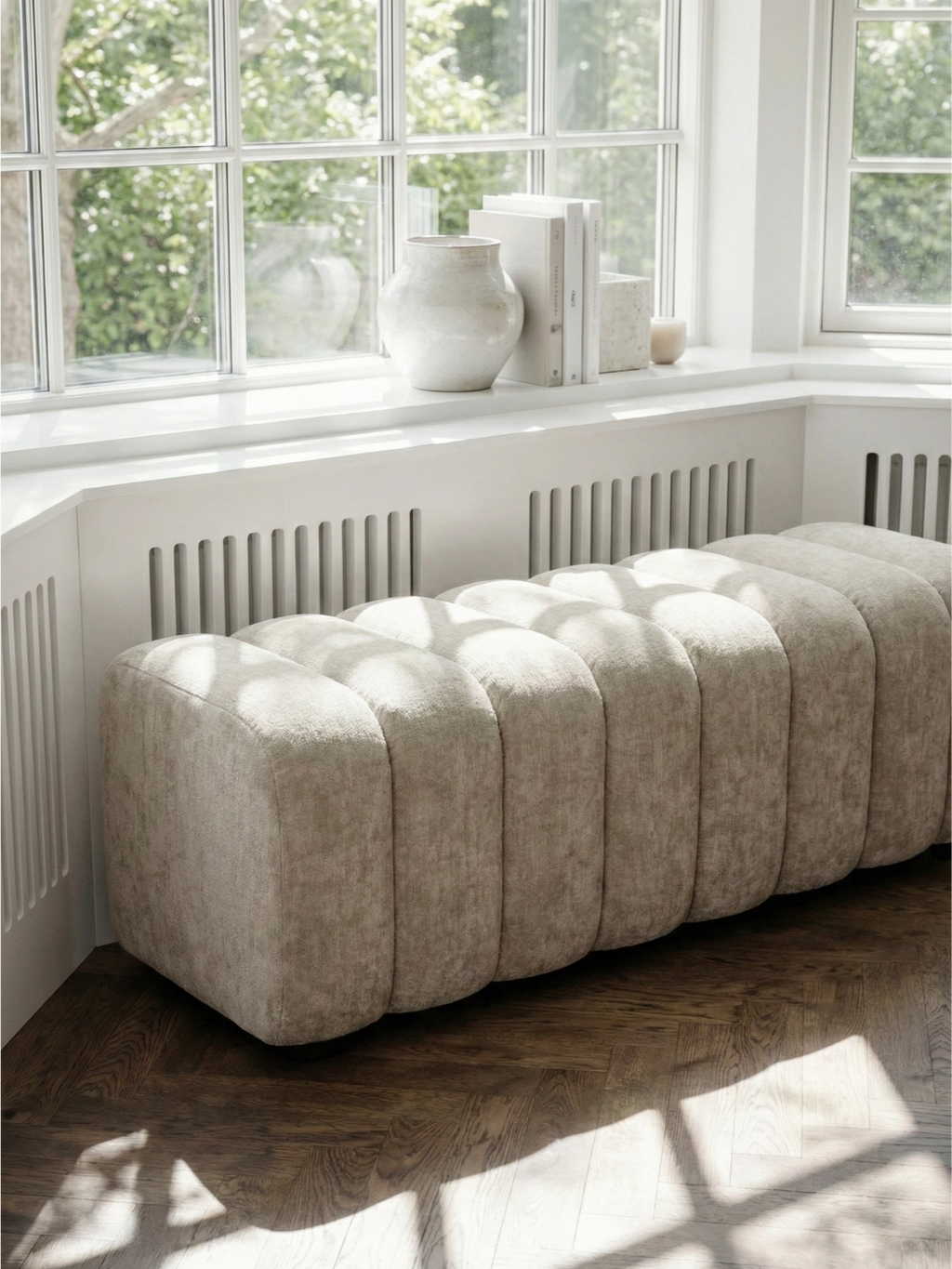 NORR11 Studio Bench. Close-up of the elegant light beige upholstered bench, featuring distinctive vertical channel tufting and a plush, inviting texture, positioned by a sunlit window.