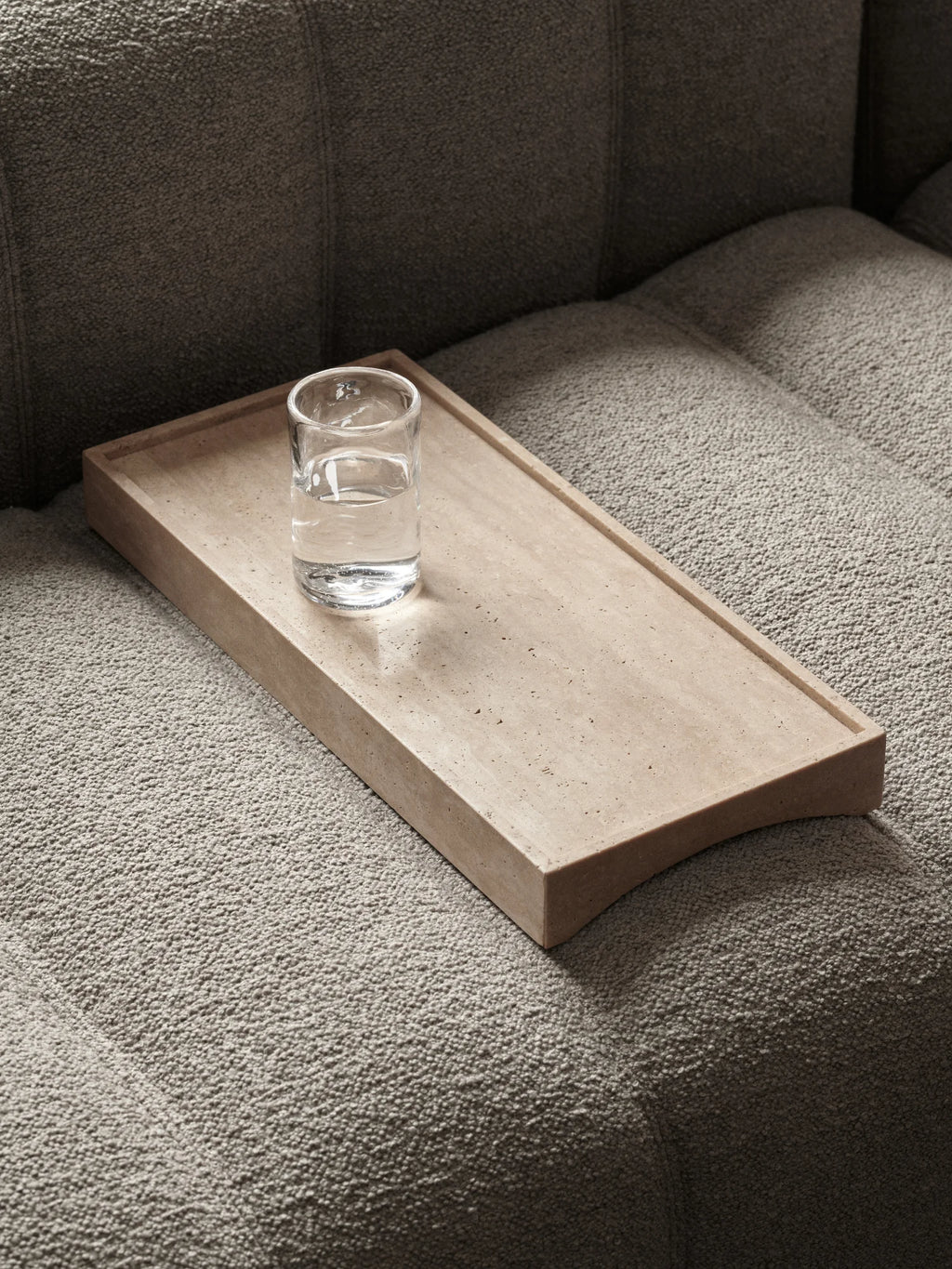 NORR11 Studio Tray, a minimalist beige travertine tray with a raw, porous texture and a subtly curved end, featuring raised edges for functional use. This organic modern natural stone tray offers a sleek, contemporary design in warm sandy tones. Ideal for sofa armrest styling, as a decorative living room tray, or a brutalist accent piece.