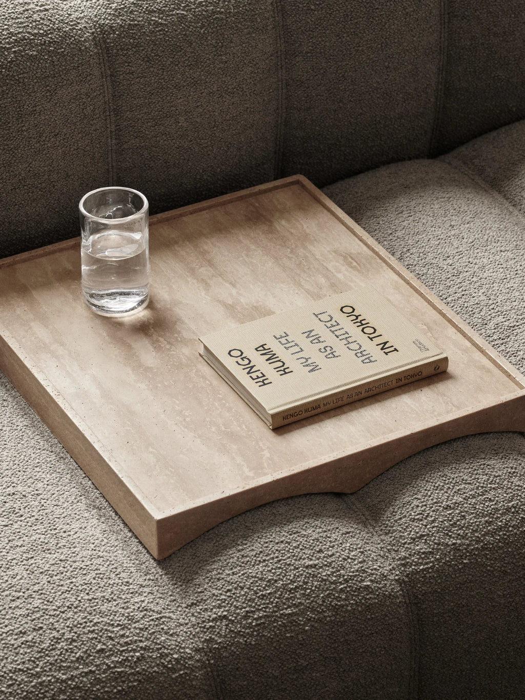 NORR11 Studio Tray, a rectangular organic modern natural travertine stone tray in a light beige finish with a distinct porous texture and elevated edges, specifically designed to fit over a sofa or couch arm. This minimalist decorative tray is ideal for contemporary living rooms, bedside tables, or Scandinavian-inspired interiors.