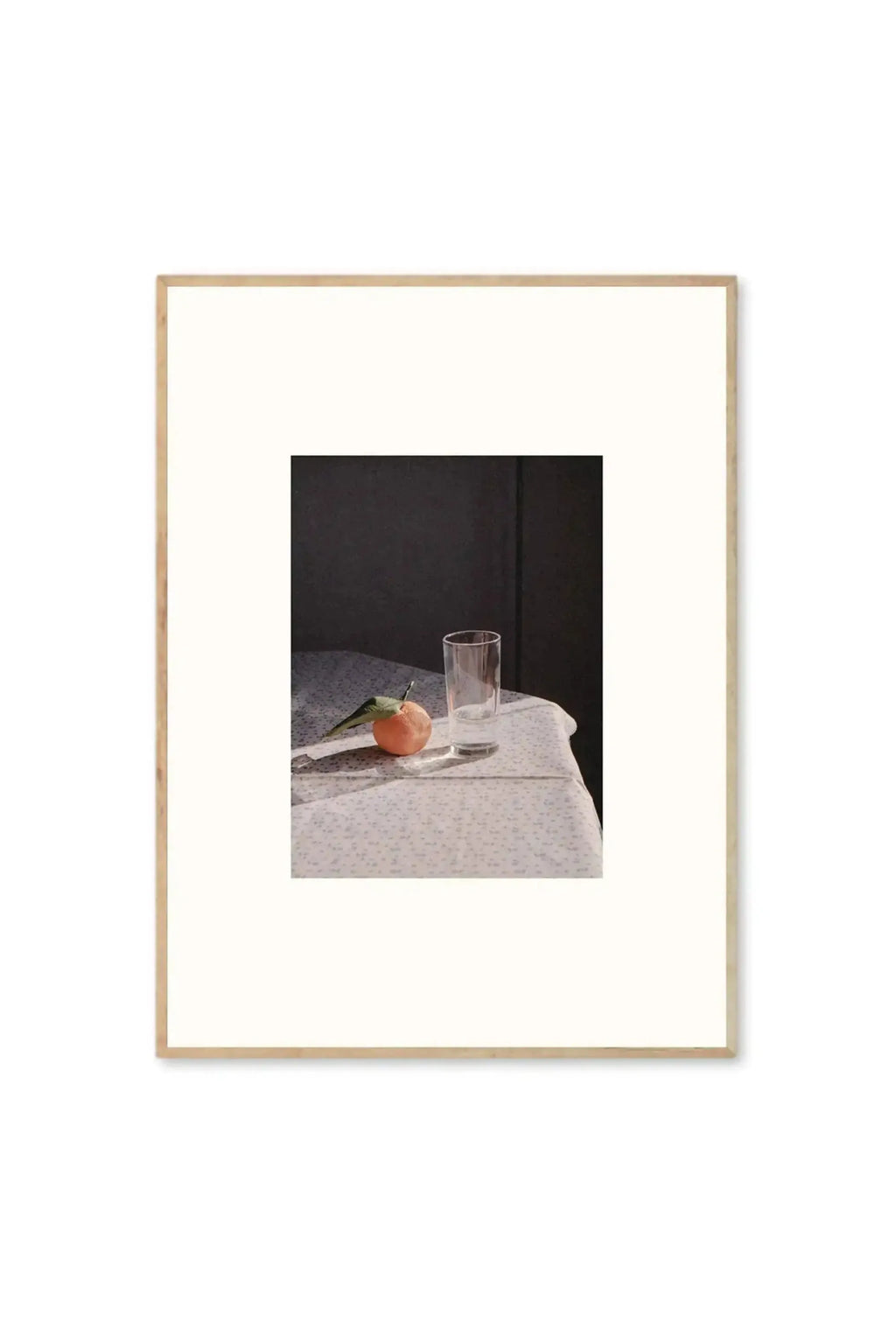 The Poster Club Sunrise - Art Print, a still life photograph featuring a single orange with a green leaf and a clear glass on a light blue patterned tablecloth, framed in natural light wood. This aesthetic wall decor poster is perfect for adding a tranquil, modern touch to any contemporary home or office.
