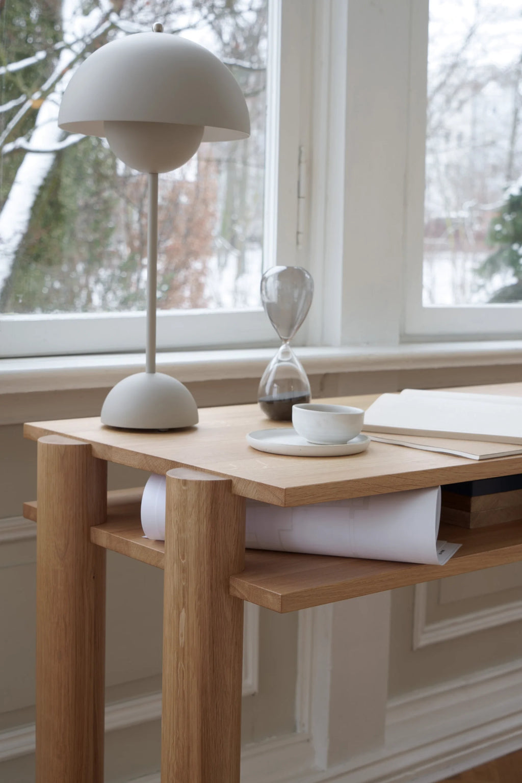 Wooden desk with a lamp, cup, and book in a room with large windows showing trees.