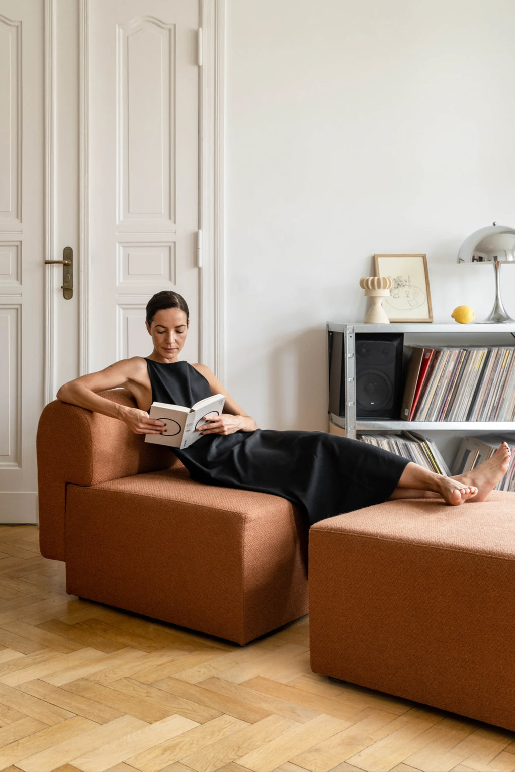 Woman reading a book on a brown armchair in a room with white walls and wooden floor.