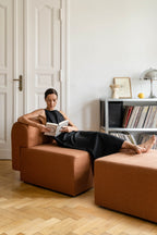 Woman reading a book on a brown armchair in a room with white walls and wooden floor.