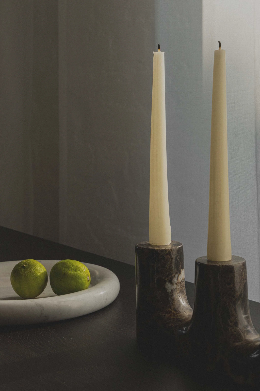 TAMO Trilo Candle Holder featuring two polished dark brown marble candle stands with cream taper candles in a lifestyle setting, with a white marble bowl and two limes in the foreground. Adds luxurious ambiance to any room.