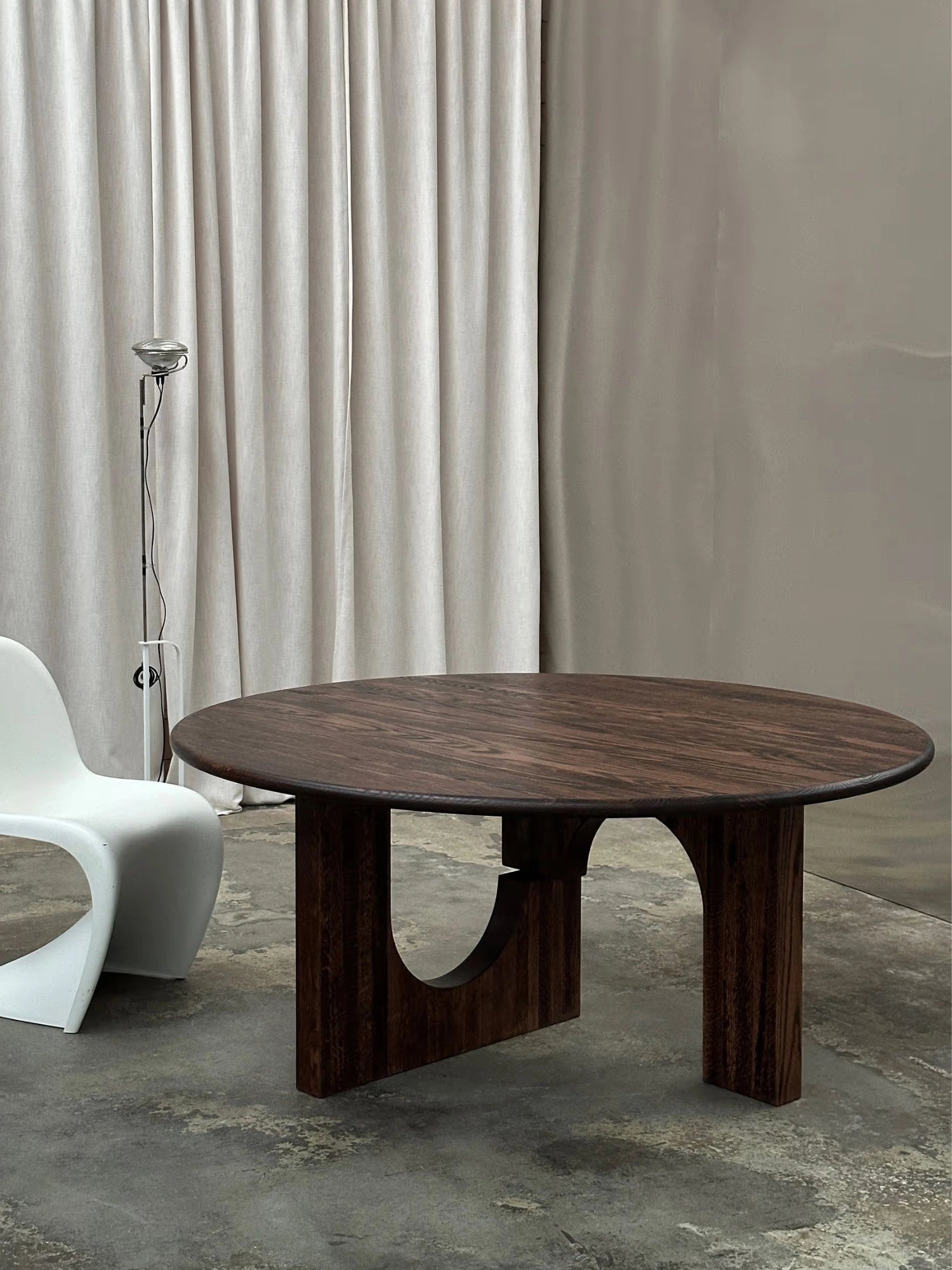 Tinted Objects Sanders Round Dining Table, a statement-making dark solid wood dining table featuring a richly grained round tabletop and a substantial sculptural base with bold, brutalist-inspired arched cutouts.