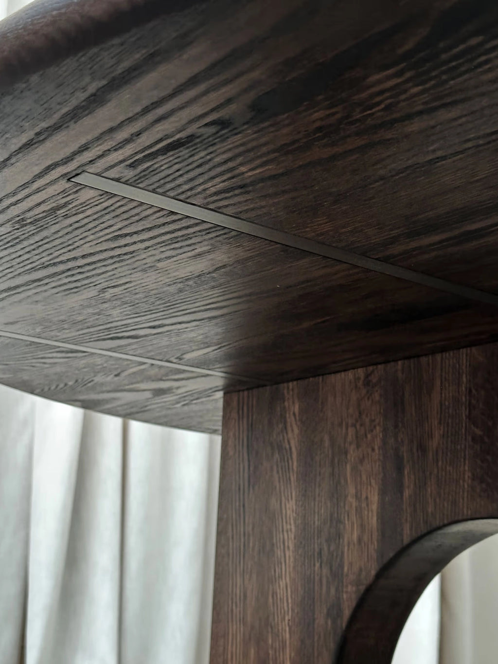 Tinted Objects Sanders Round Dining Table, featuring a rich, dark brown oak wood grain with subtle dark gray metallic inlays near the table's edge and along the pedestal base. This elegant, modern dining table is ideal for contemporary homes and design-conscious individuals seeking a stylish and durable centerpiece.