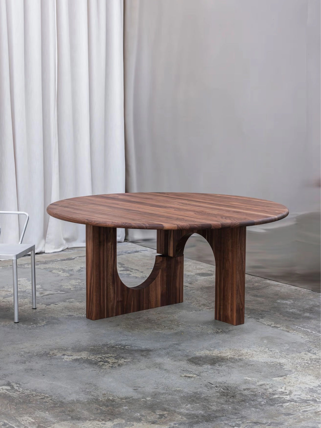 Tinted Objects Sanders Round Dining Table, a premium solid dark walnut wood table with a smooth, round top and a unique sculptural base featuring two arched cutouts. This modern and elegant dining table is perfect for contemporary homes seeking stylish, durable furniture.