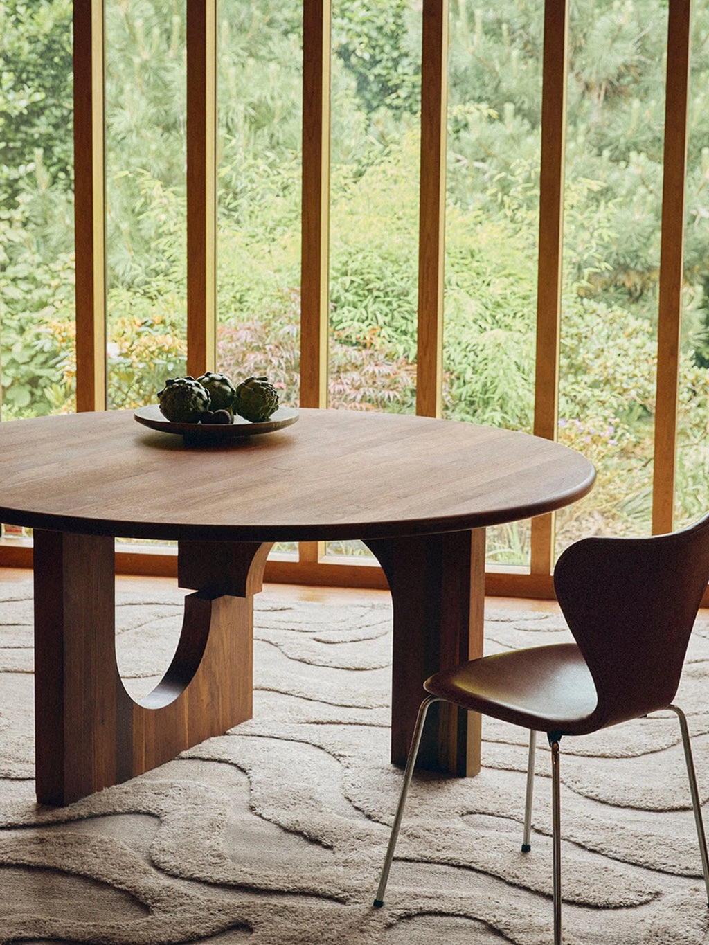 Tinted Objects Sanders Round Dining Table, a stylish dark brown wooden dining table with a unique curved pedestal base and a smooth round top. This modern dining table is perfect for elegant dining rooms and contemporary living spaces.