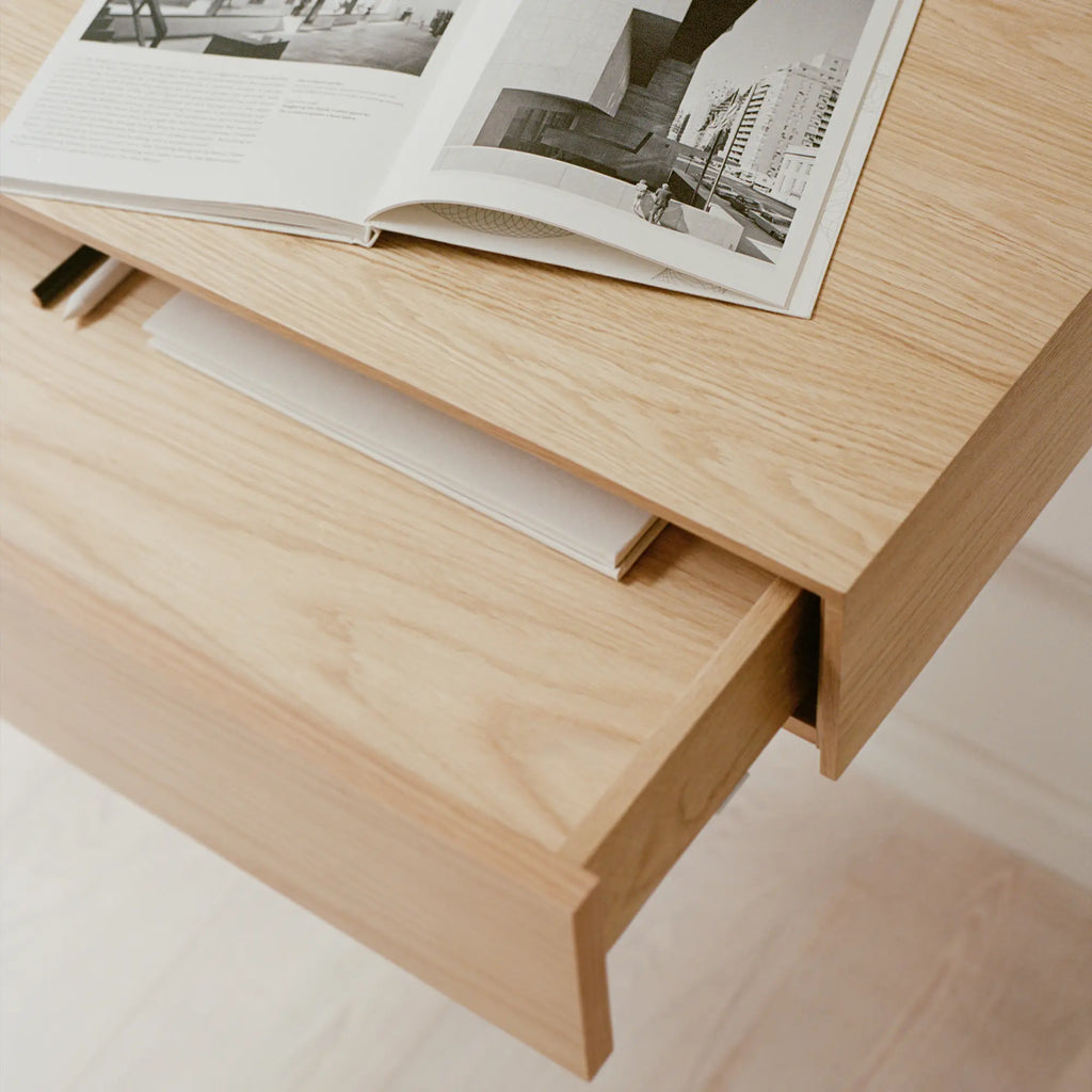 New Works Tana Wall Mounted Desk, a modern minimalist wall-mounted desk crafted from light natural solid oak with visible wood grain. It features a clean rectangular silhouette and a convenient pull-out storage drawer, creating a sleek floating design. Ideal as a space-saving home office desk, a Scandinavian-inspired console, or an elegant entryway table for contemporary interiors.