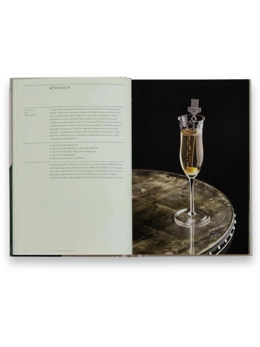 Book The Connaught Bar, an elegant cocktail recipe book open to a "Benvenuti" page with a detailed recipe for a sparkling champagne cocktail and an enticing image of the finished drink in a flute glass with a pineapple garnish and a branded stirrer, all set on a dark, textured table. This premium book is perfect for home mixologists and cocktail enthusiasts seeking sophisticated drink recipes.