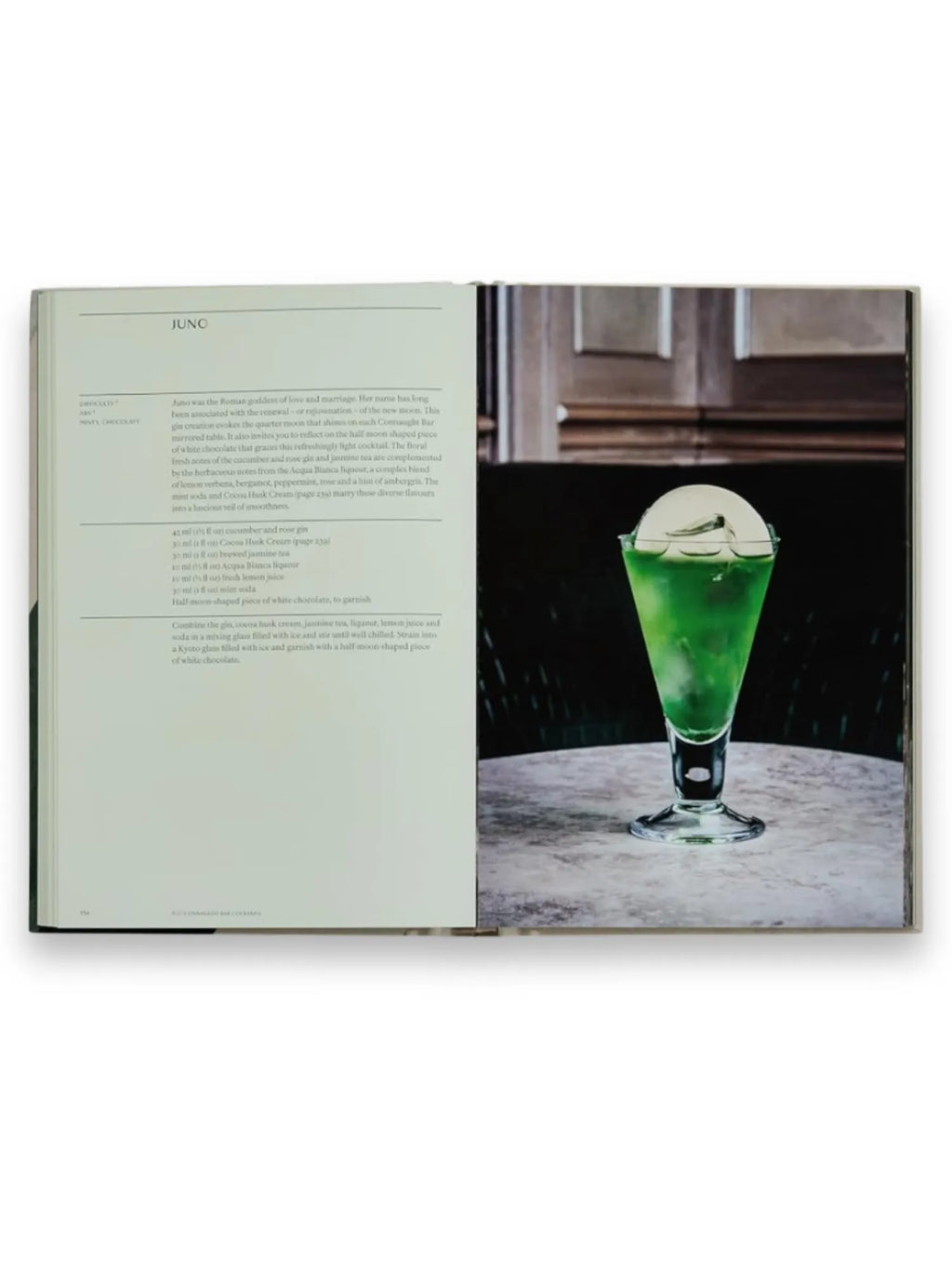Book The Connaught Bar, an open cookbook displaying a vibrant green cocktail recipe with descriptive text and a full-page photo of a tall, clear glass filled with a verdant drink, topped with white foam and a half-moon white chocolate garnish, set against a blurred luxurious bar background. This premium cocktail recipe book is perfect for mixology enthusiasts and home bartenders seeking elegant, sophisticated drink ideas.