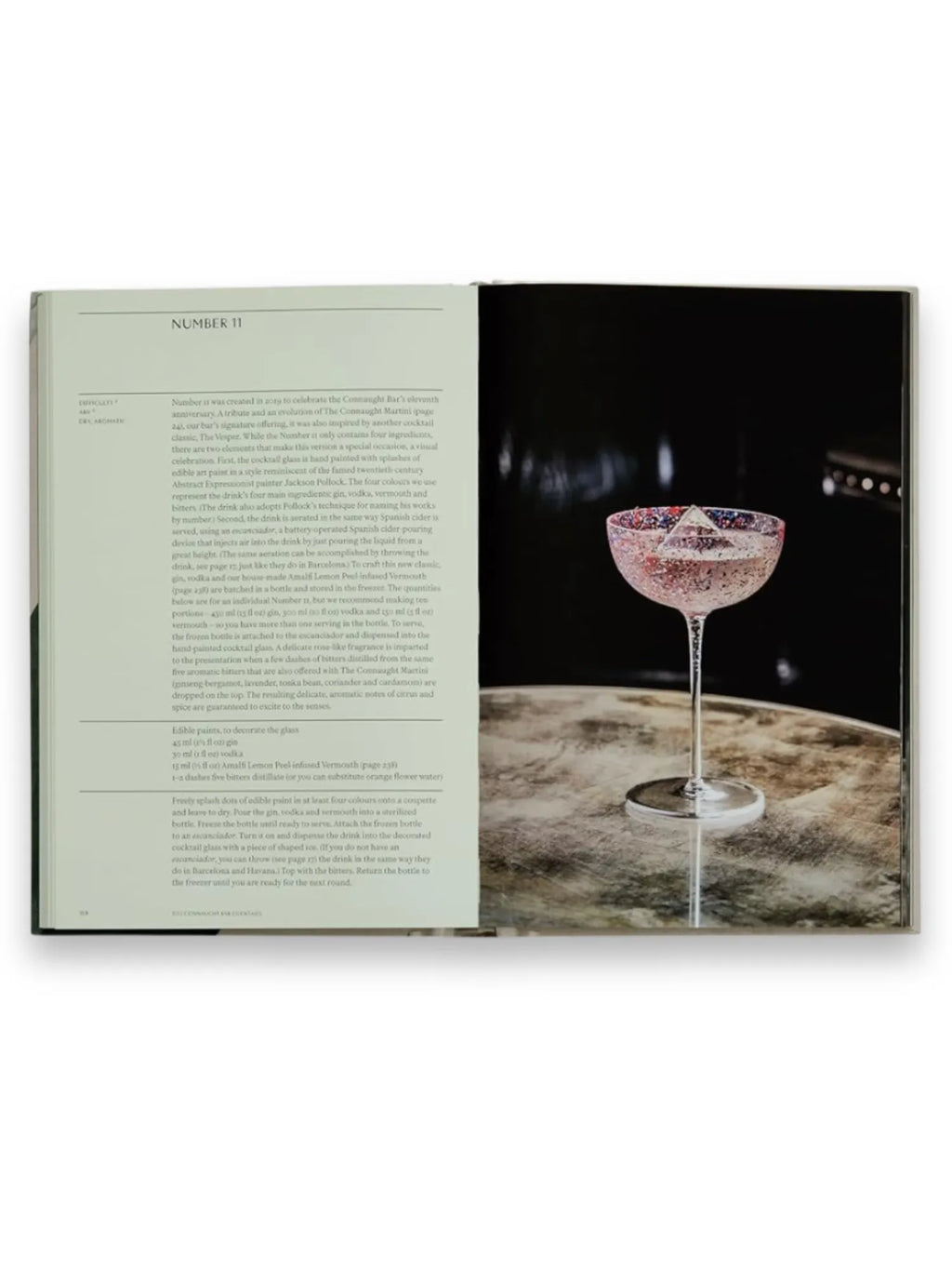 Book The Connaught Bar, an open cookbook displaying a vibrant recipe for "Number 11," featuring a stunning cocktail in a hand-painted coupe glass with a pyramid ice cube on a textured marble-like surface. This luxurious cocktail recipe book is perfect for mixology enthusiasts and home bartenders seeking sophisticated drink creations.