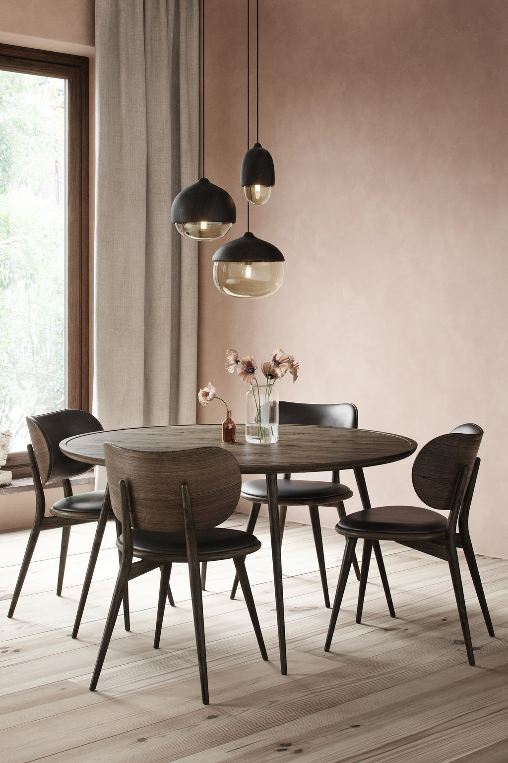 Mater The Dining Chair, a set of four dark wood dining chairs with black leather seats, arranged around a round wooden dining table in a cozy dining room with a soft pink wall and modern pendant lights, creating an inviting atmosphere.