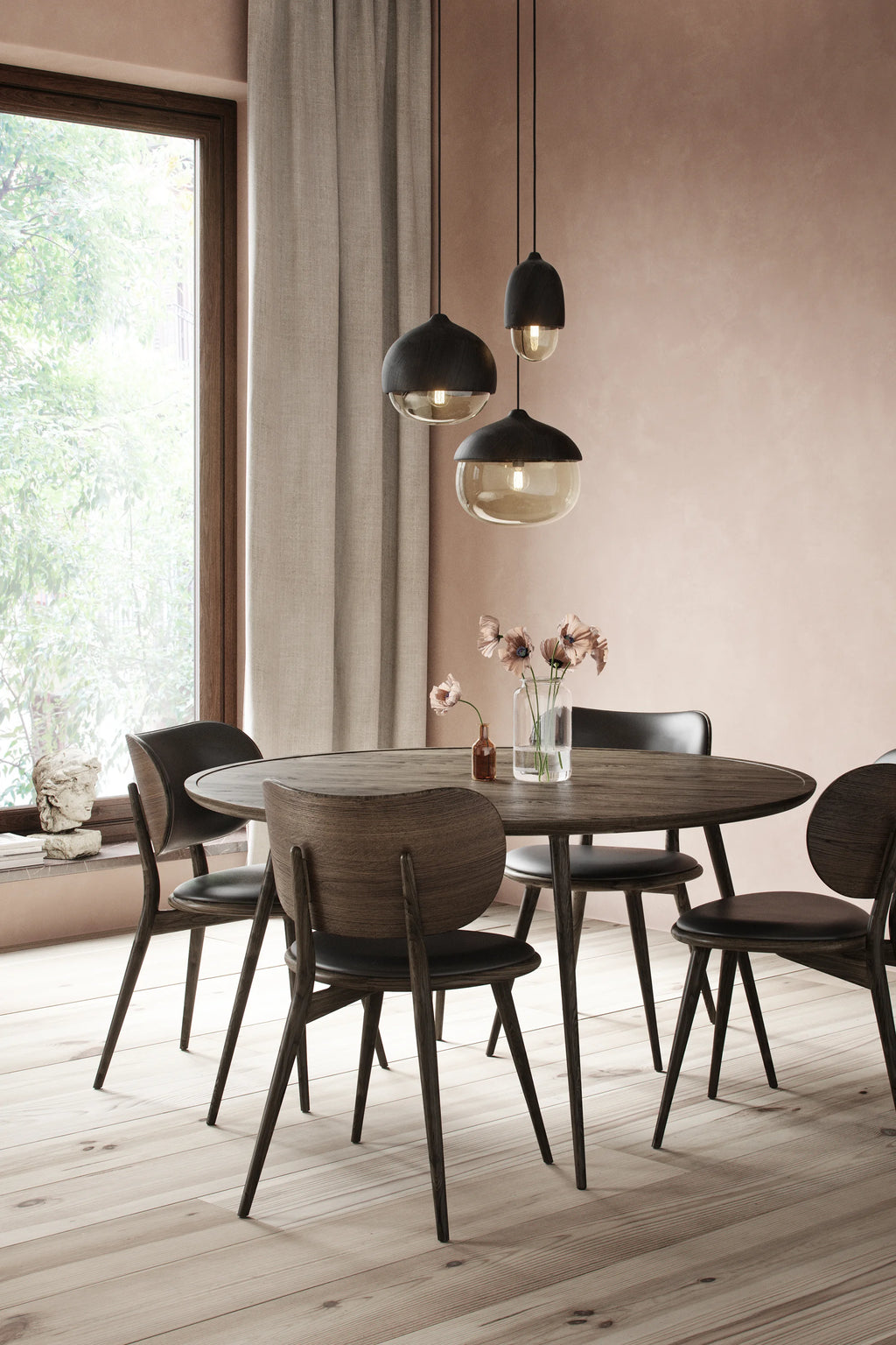 Mater Accent Round Dining Table, in a rich, dark wood finish, surrounded by four matching dining chairs with black seats, set in an elegant dining room with a soft pink wall, large window, and hanging pendant lights.