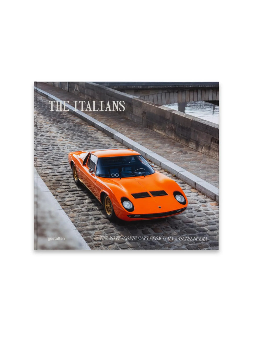 Book The Italians - Beautiful Machines, a premium hardcover book featuring a vibrant orange vintage Italian sports car with gold wheels on a cobblestone street, with a stone bridge and water in the background. This stylish coffee table book is perfect for car enthusiasts and collectors of automotive history and design.