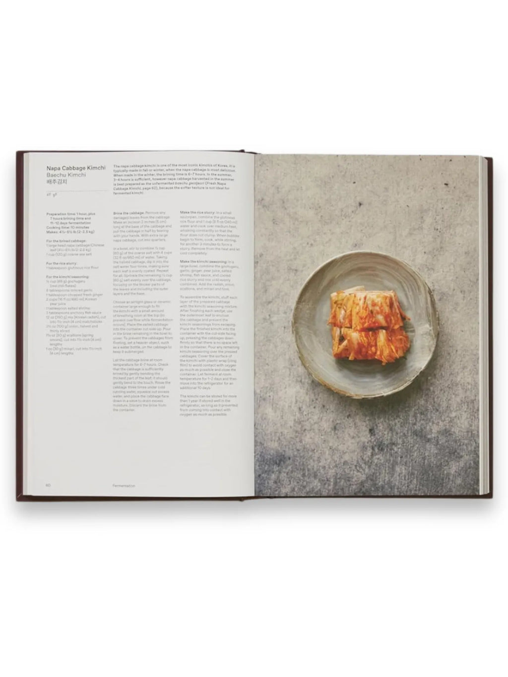 Book The Korean Cookbook, an open hardcover cookbook showcasing a detailed "Napa Cabbage Kimchi" recipe with ingredients and instructions on the left page. The right page features a beautiful, high-resolution food photograph of vibrant red kimchi served in a rustic grey ceramic bowl, centered on a textured, light grey surface. This essential culinary guide is perfect for home cooks and food enthusiasts eager to master authentic Korean cuisine.