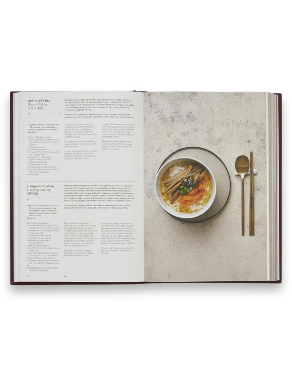 Book The Korean Cookbook, an open hardcover cookbook displaying a recipe for "Acorn Jelly Bap" and a photograph of a beautifully plated Korean dish in a round bowl with gold-toned flatware. This essential culinary guide is perfect for home cooks and food enthusiasts exploring authentic Korean cuisine.