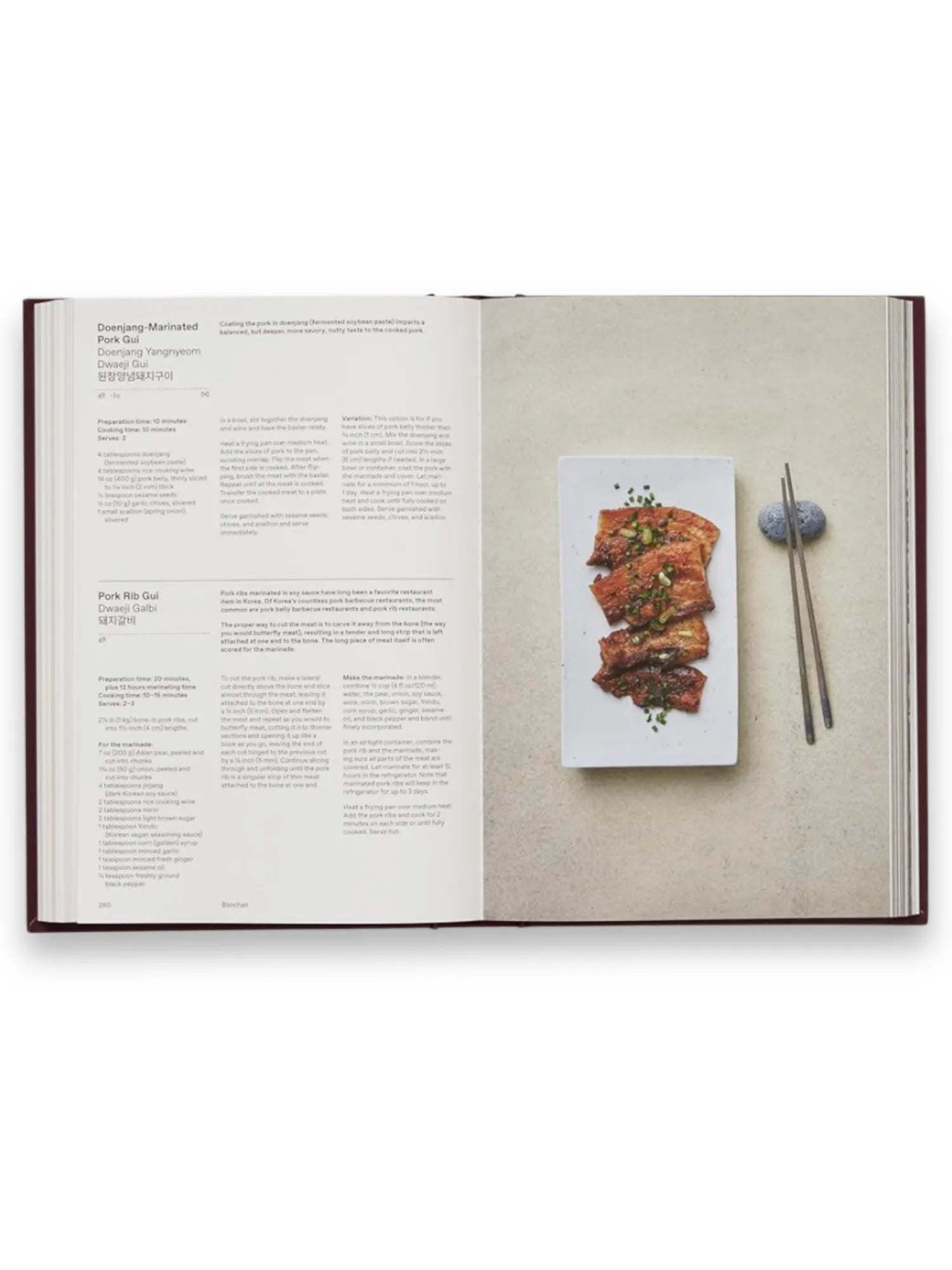 Book The Korean Cookbook, an open hardcover cookbook showcasing a detailed recipe for "Doenjang-Marinated Pork Gui" with a vibrant, high-quality photograph of the dish on a white rectangular plate next to elegant chopsticks on a stone rest. This essential culinary guide is perfect for home cooks eager to explore authentic Korean cuisine and cooking techniques.