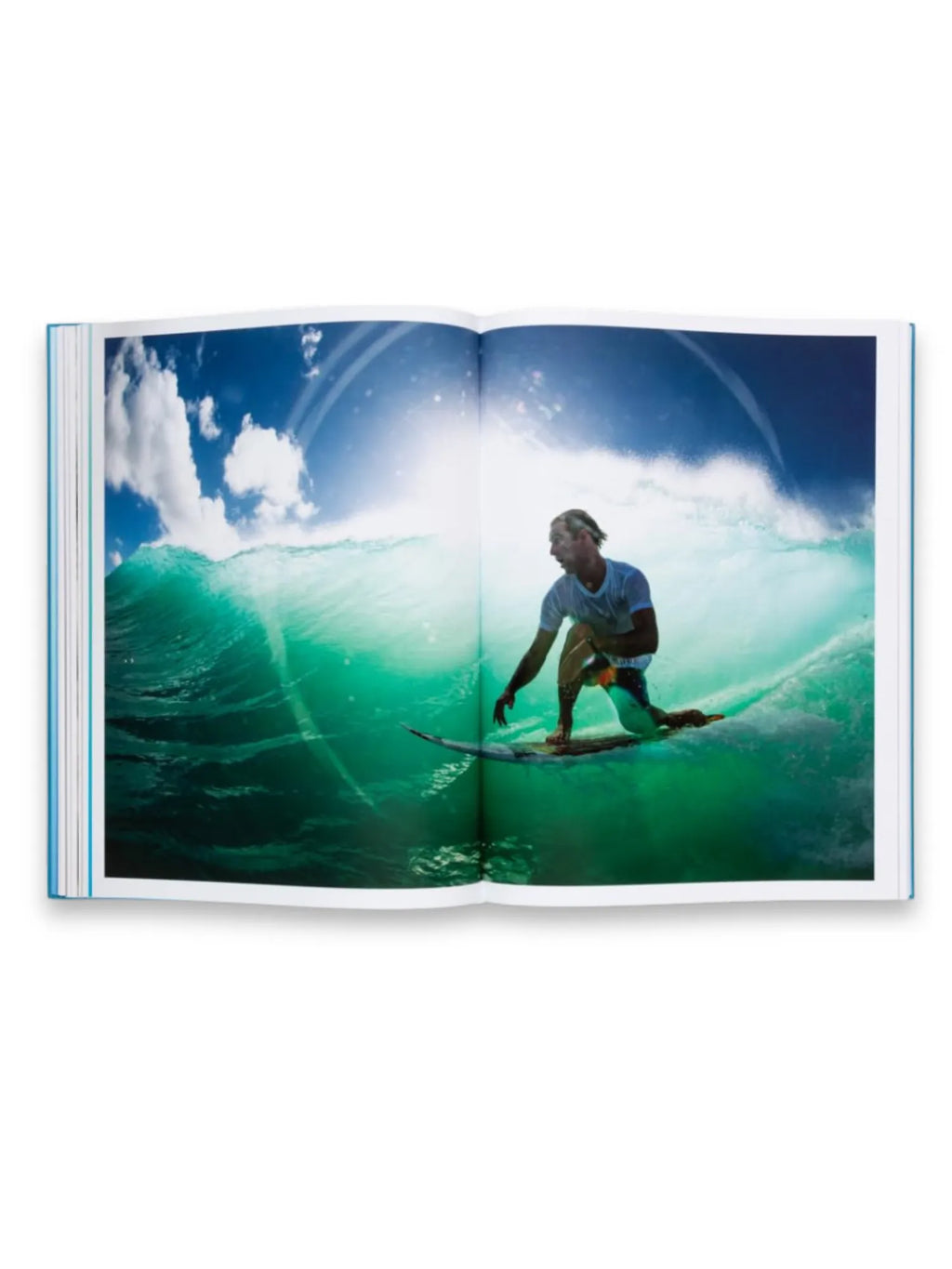 Book The Oceans, an open hardcover book displaying a vibrant photograph of a surfer riding a large, turquoise-green ocean wave under a bright blue sky with white clouds. This visually stunning photography book is perfect for surf enthusiasts, travelers, and ocean lovers seeking inspiring imagery.