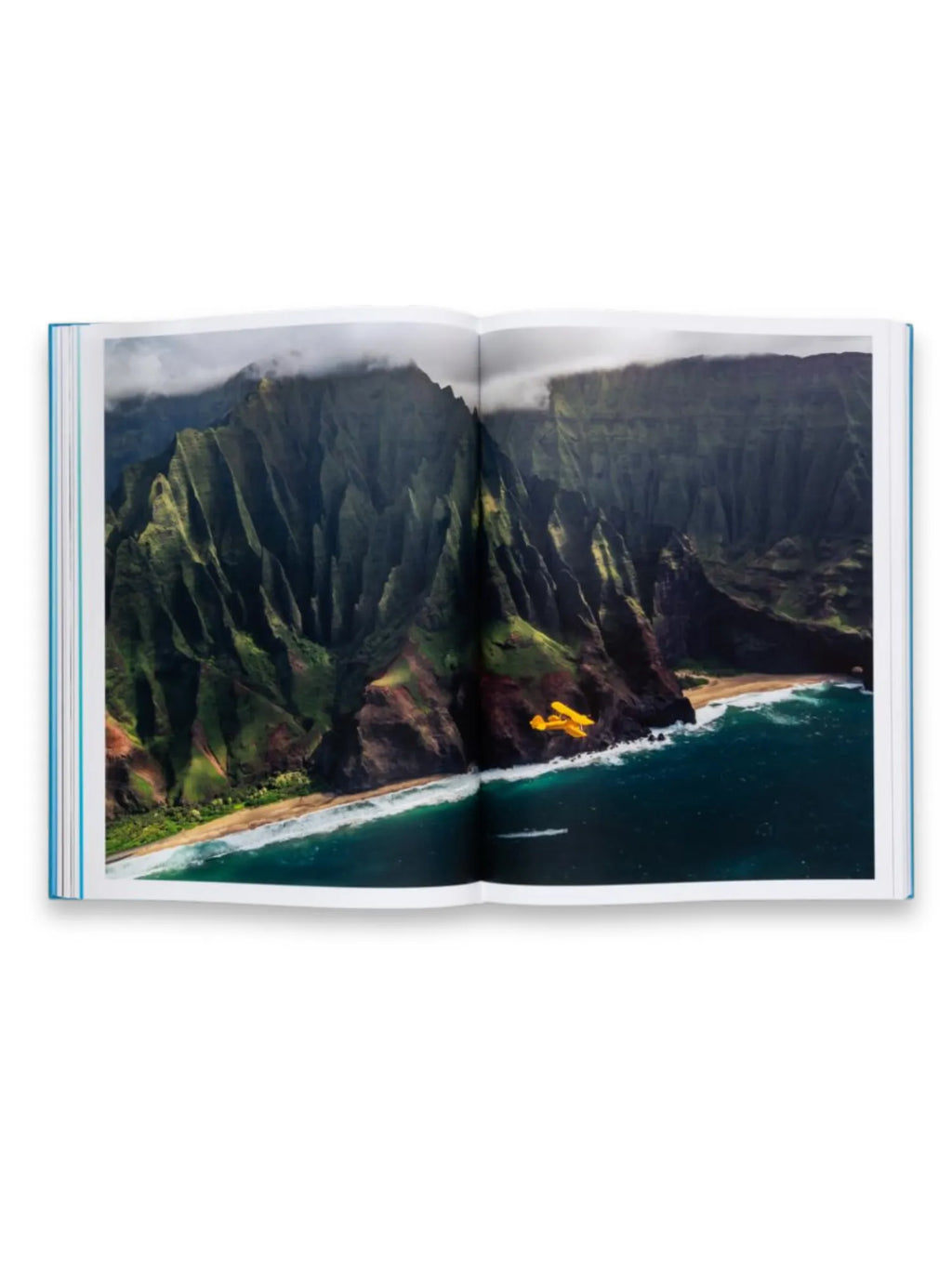 Book The Oceans, an open book displaying a vibrant aerial photo of a rugged green coastline with steep cliffs, a sandy beach, and turquoise ocean waves, featuring a bright yellow biplane flying low over the water. This captivating photography book is perfect for travel enthusiasts and nature lovers seeking stunning visual inspiration.