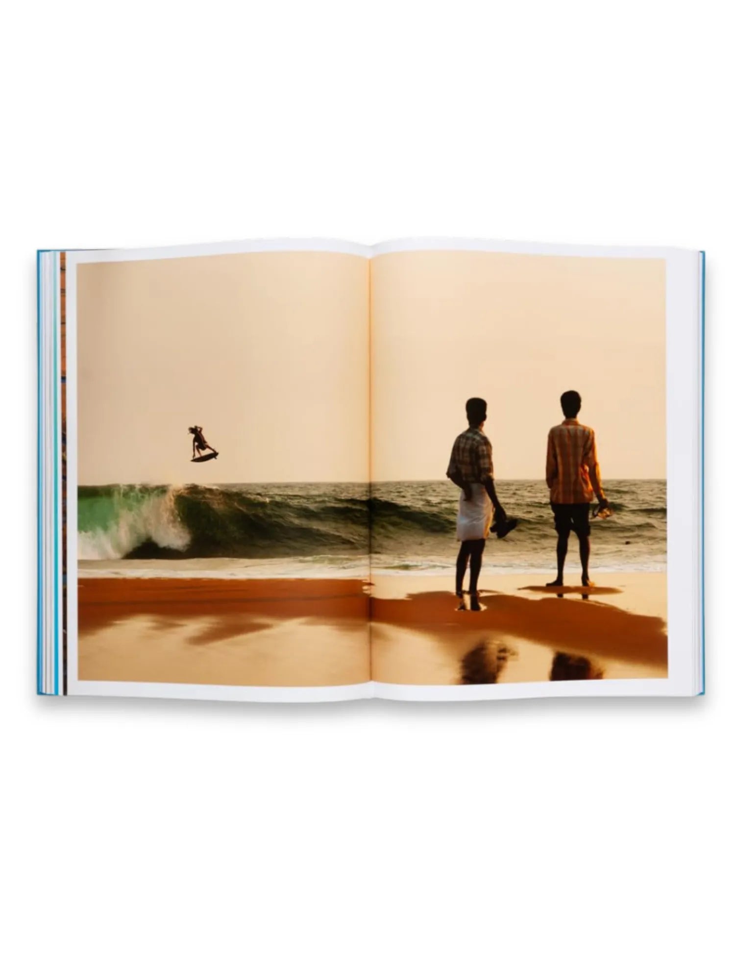 Book The Oceans, an open book displaying a vibrant photographic spread of a beach scene at sunset with a surfer airborne over a turquoise wave and two men standing on the golden sand. This premium photography book offers stunning visual narratives for art lovers and travel enthusiasts.