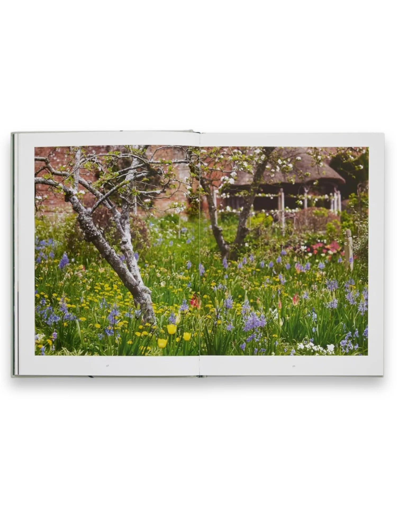 Book The Tulip Garden, an open hardcover book displaying a vibrant, full-page photograph of a lush garden scene with blooming yellow tulips, purple hyacinths, and white flowers amidst green foliage, with a rustic thatched-roof gazebo in the background. This beautiful coffee table book is perfect for garden enthusiasts and home decor lovers seeking nature-inspired inspiration.