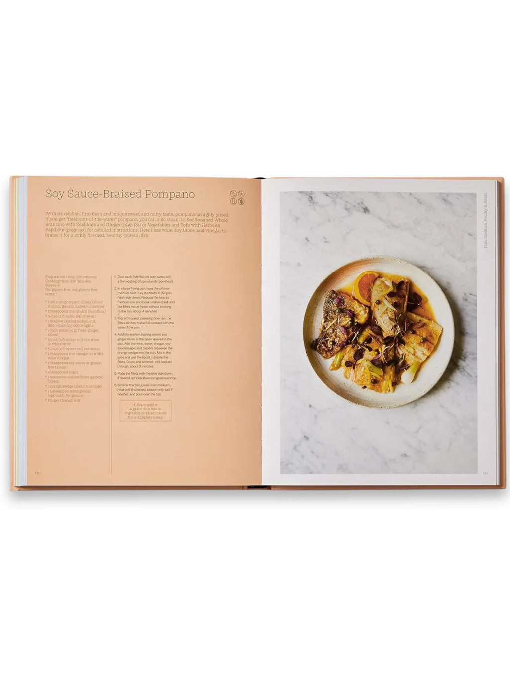 Book The Wellness Principles: Cooking for a Healthy Life, an open hardcover cookbook showcasing a recipe for Soy Sauce-Braised Pompano with detailed instructions and a vibrant, beautifully plated dish on a speckled white ceramic plate against a white marble background. This essential healthy cooking guide offers delicious, wholesome recipes for home cooks focused on nutritious meals.