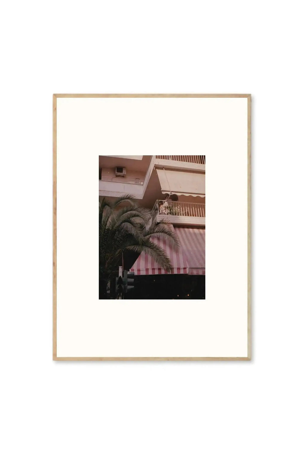 The Poster Club The Corner We Both Loved - Art Print, a framed photograph featuring a light pink building with a striped pink and white awning, and a lush green palm tree in the foreground. This stylish wall art adds a touch of modern elegance to any contemporary home decor.
