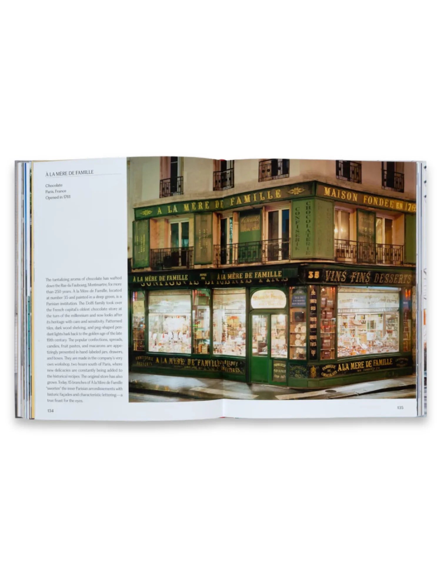 Book Think Big Shop Small, an open hardcover book displaying a vibrant photograph of "À la Mère de Famille", a historic Parisian chocolate shop with a deep green facade, intricate gold lettering, and beautifully lit display windows filled with confections. This appealing book offers a delightful journey into the world of small businesses and unique establishments, perfect for entrepreneurs, history buffs, and travel enthusiasts alike.
