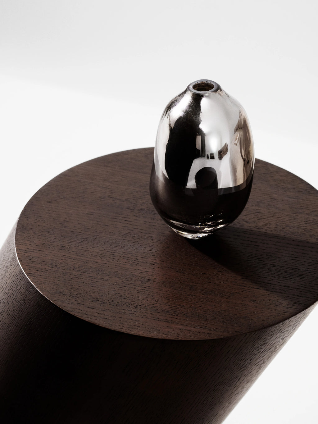 NORR11 Tilt Side Table, a modern minimalist side table crafted from dark stained oak with a striking cylindrical base and a unique tilted round top. Its rich, deep brown finish highlights the subtle wood grain, creating a sculptural and contemporary accent piece.