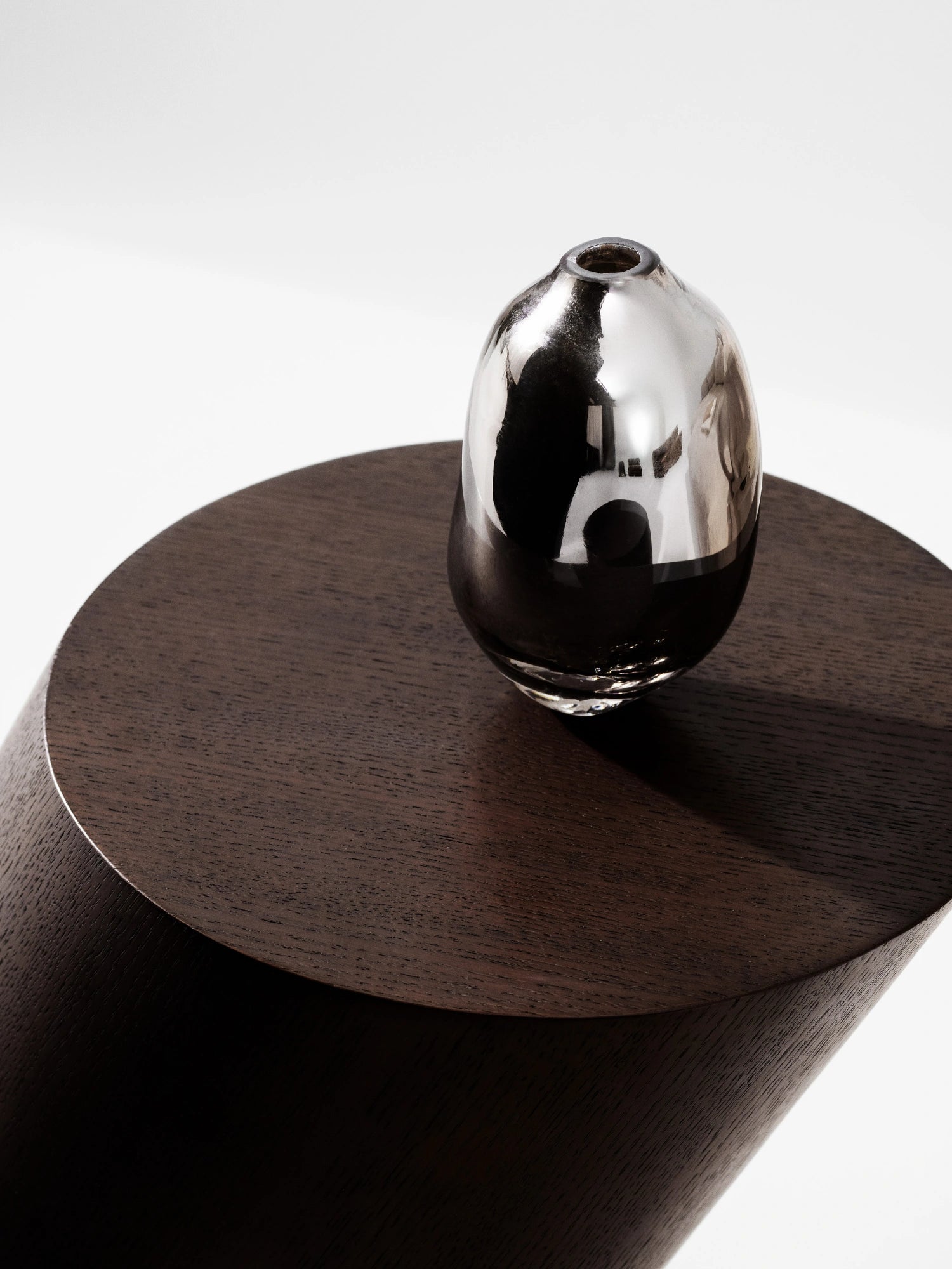 NORR11 Tilt Side Table, a modern minimalist side table crafted from dark stained oak with a striking cylindrical base and a unique tilted round top. Its rich, deep brown finish highlights the subtle wood grain, creating a sculptural and contemporary accent piece.