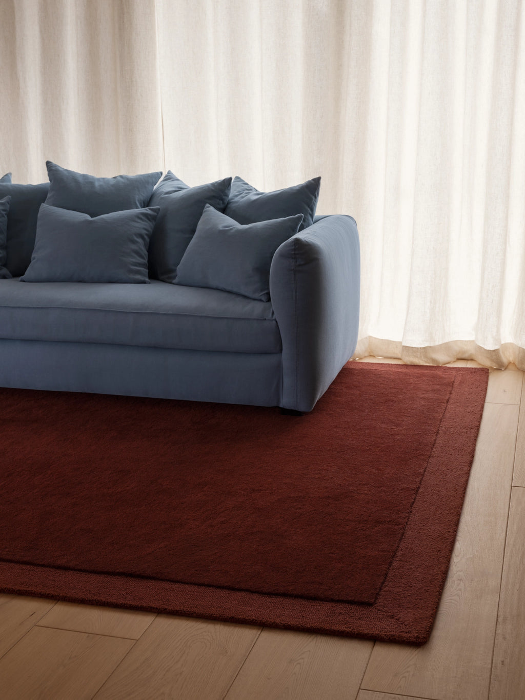 Tinted Objects Louis Wool Rug, a luxurious deep red area rug with a subtle border detail, resting on light wooden flooring. This premium natural wool rug is perfect for adding warmth and sophisticated style to any living room or bedroom.