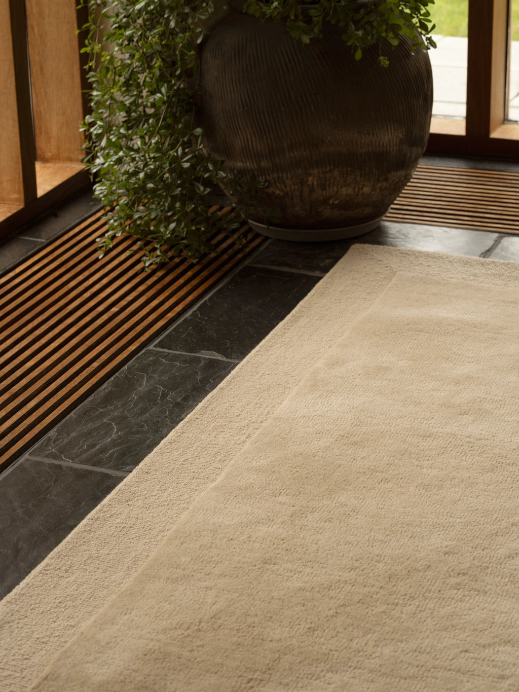 Tinted Objects Louis Wool Rug, a plush, luxurious cream-colored area rug with a subtle border detail, shown laid on dark slate tile flooring. This elegant, soft wool rug is perfect for adding warmth and sophisticated style to any contemporary living space or bedroom.