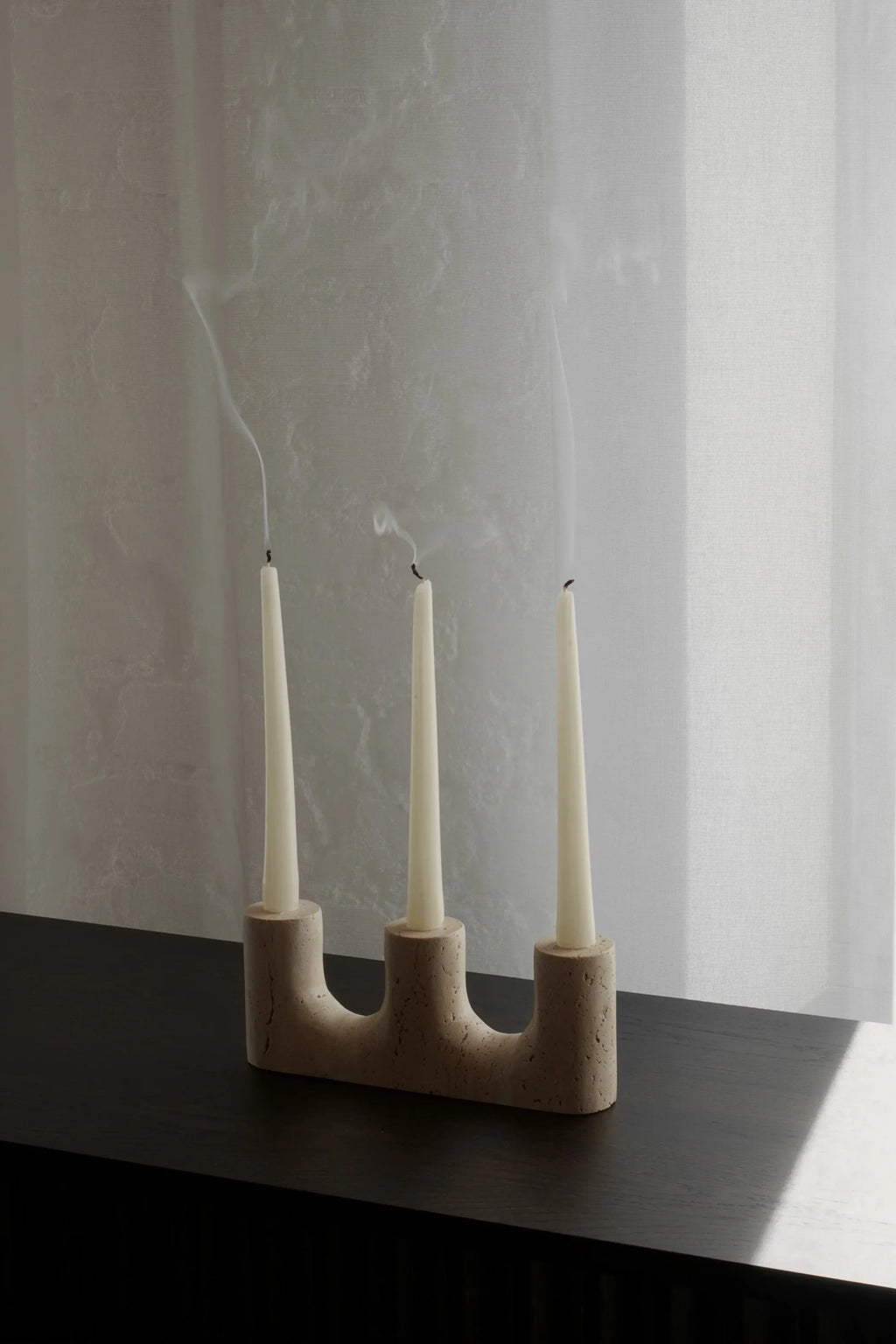 TAMO Trilo Candle Holder, a modern beige concrete candelabra holding three white taper candles with wisps of smoke, on a dark wooden surface. Perfect for minimalist home decor.