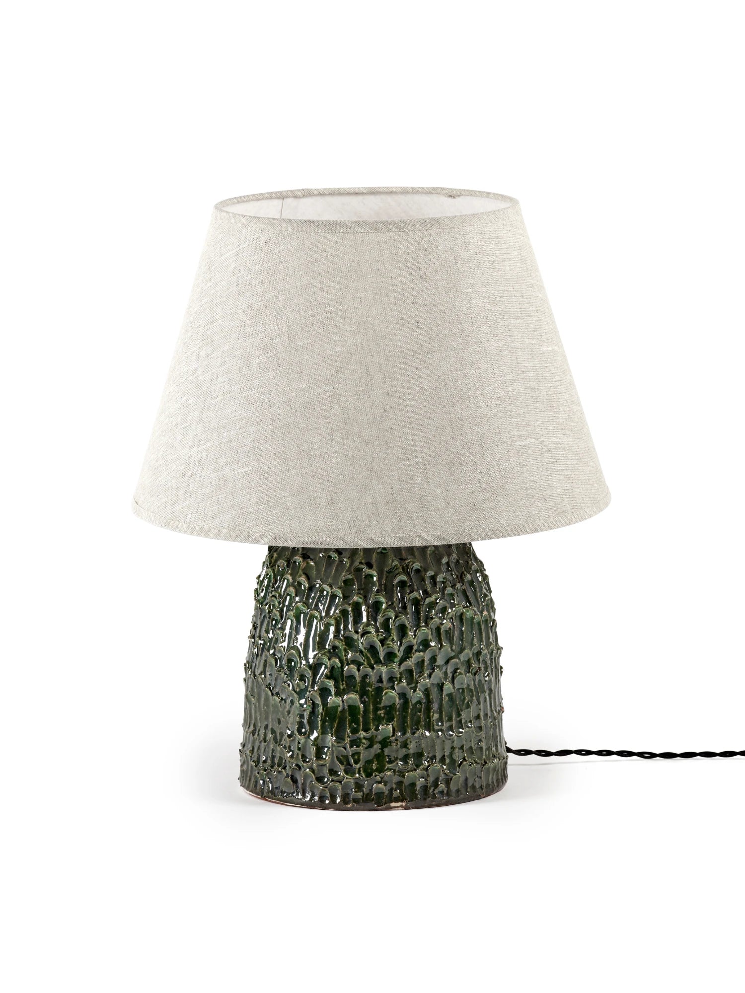 A medium-sized Tronco ceramic table lamp with a dark green, highly textured, glossy base and a conical, neutral white/beige fabric lampshade. The lamp is photographed on a white background with a black electrical cord extending to the right.